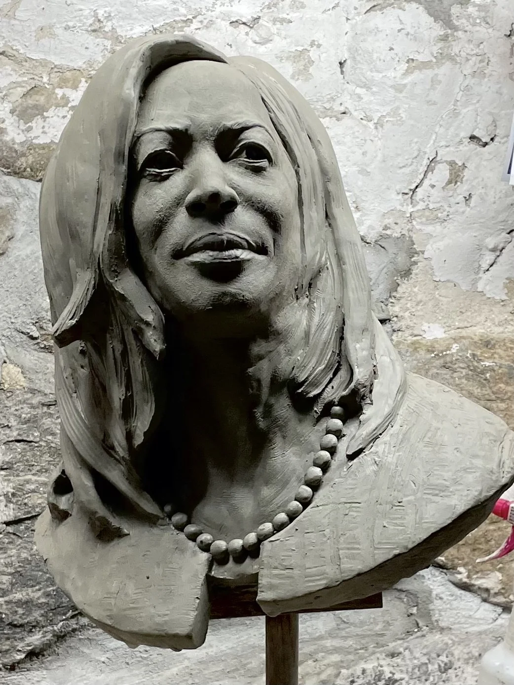 A detailed stone bust sculpture of a woman with long hair, wearing a string of beads around her neck, set against a rough stone wall.