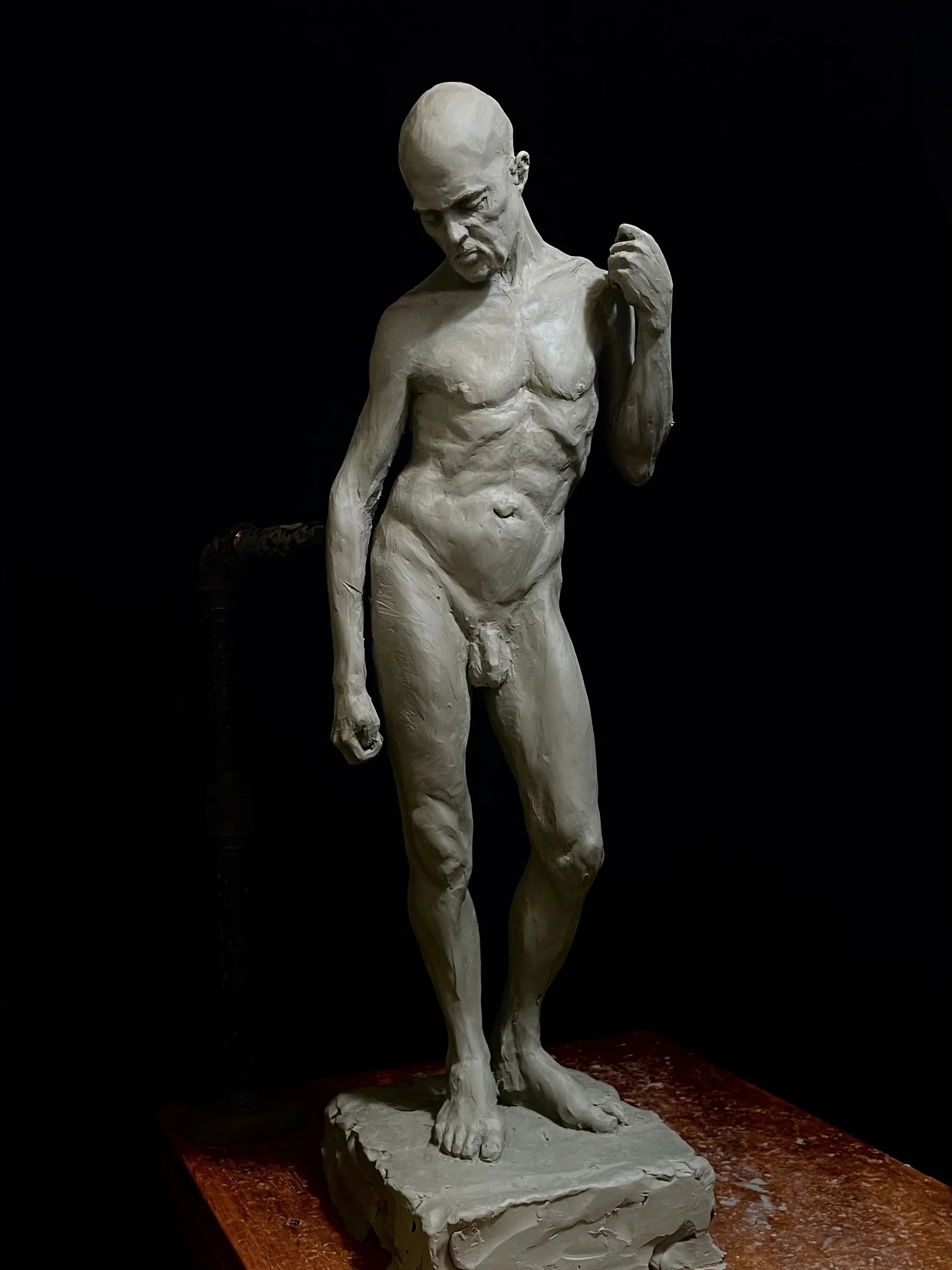 Clay sculpture of a nude man with a bald head, standing on a platform, looking downward with a contemplative expression, one arm bent holding a cloth or object near his shoulder, set against a dark background.