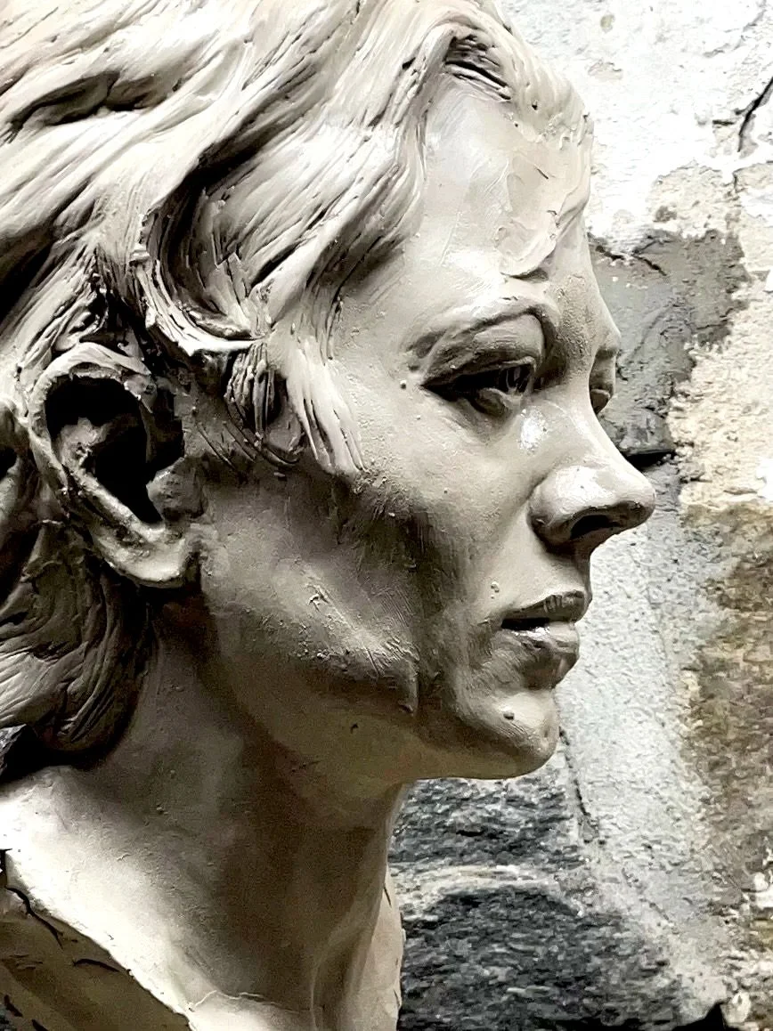 A detailed sculpture of a woman's face in profile view, made of clay or similar material, with textured hair and realistic facial features.
