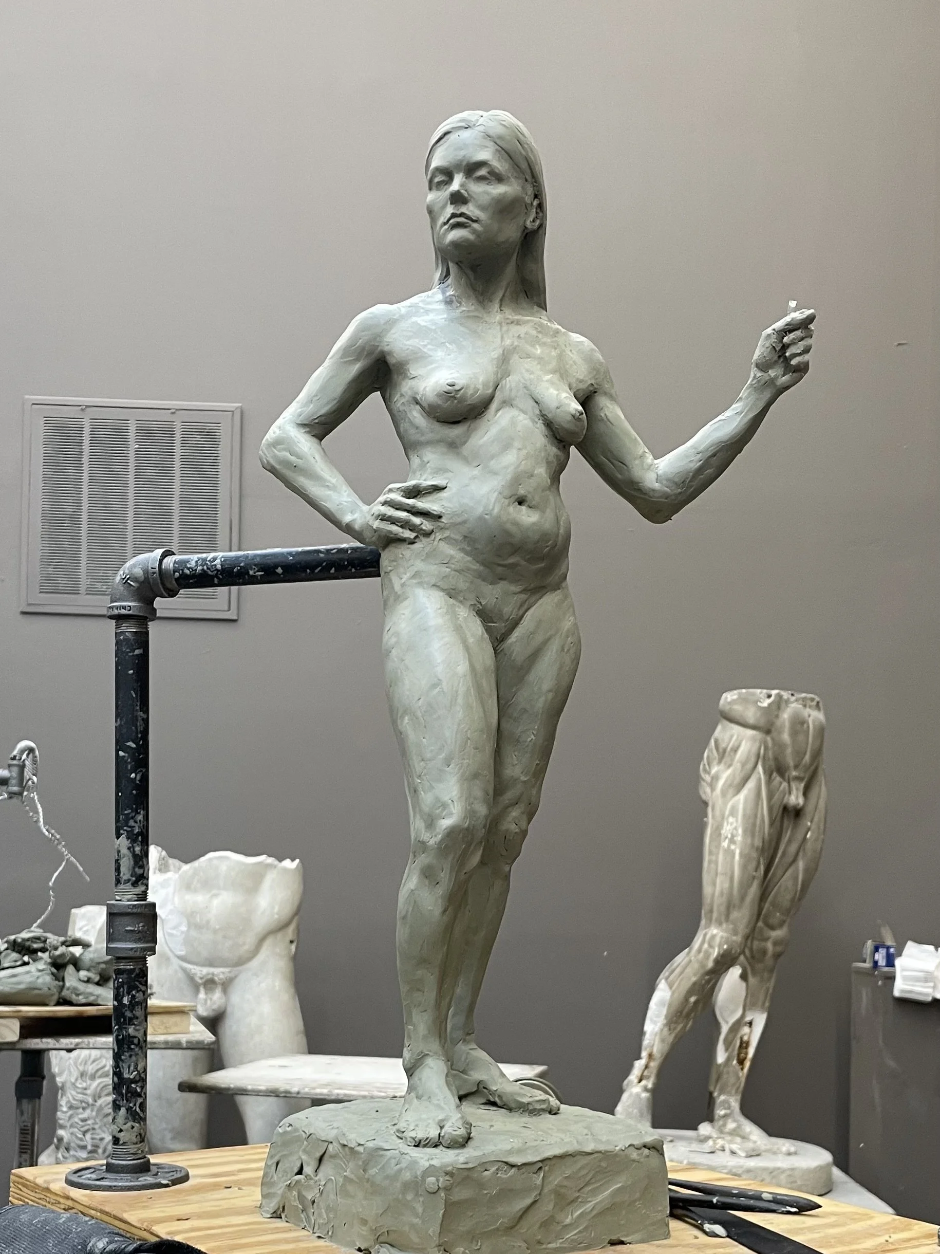 A clay sculpture of a woman with one hand on her hip and the other raised, in the process of being carved, on a wooden worktable surrounded by sculpting tools and other sculptures in progress.