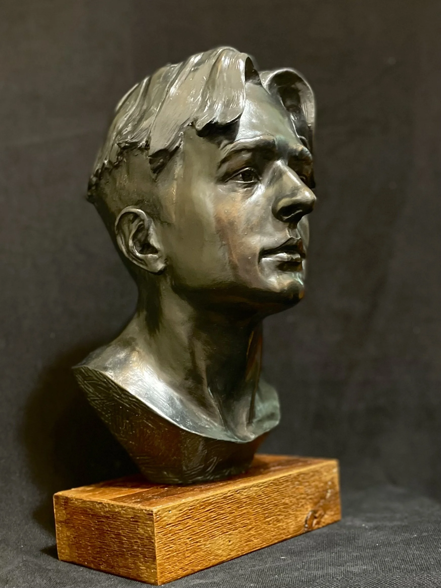 Bronze bust sculpture of a young man's head with short hair mounted on a wooden block against a black background.
