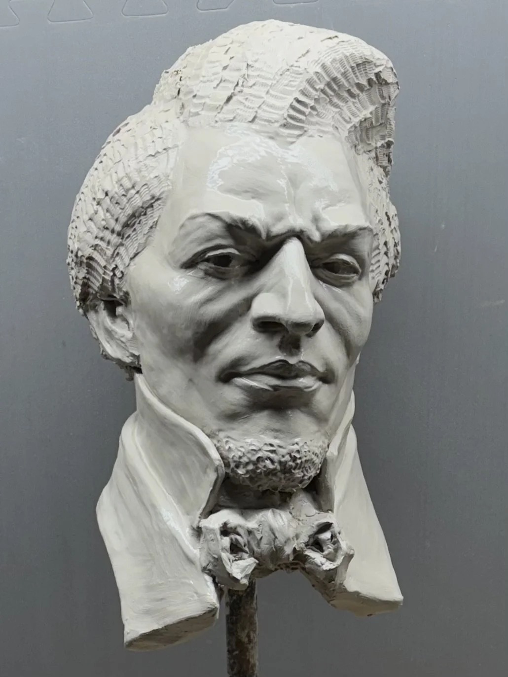 A clay sculpture bust of Frederick Douglass with stylized hair, a bow tie, and a confident expression, mounted on a stand.