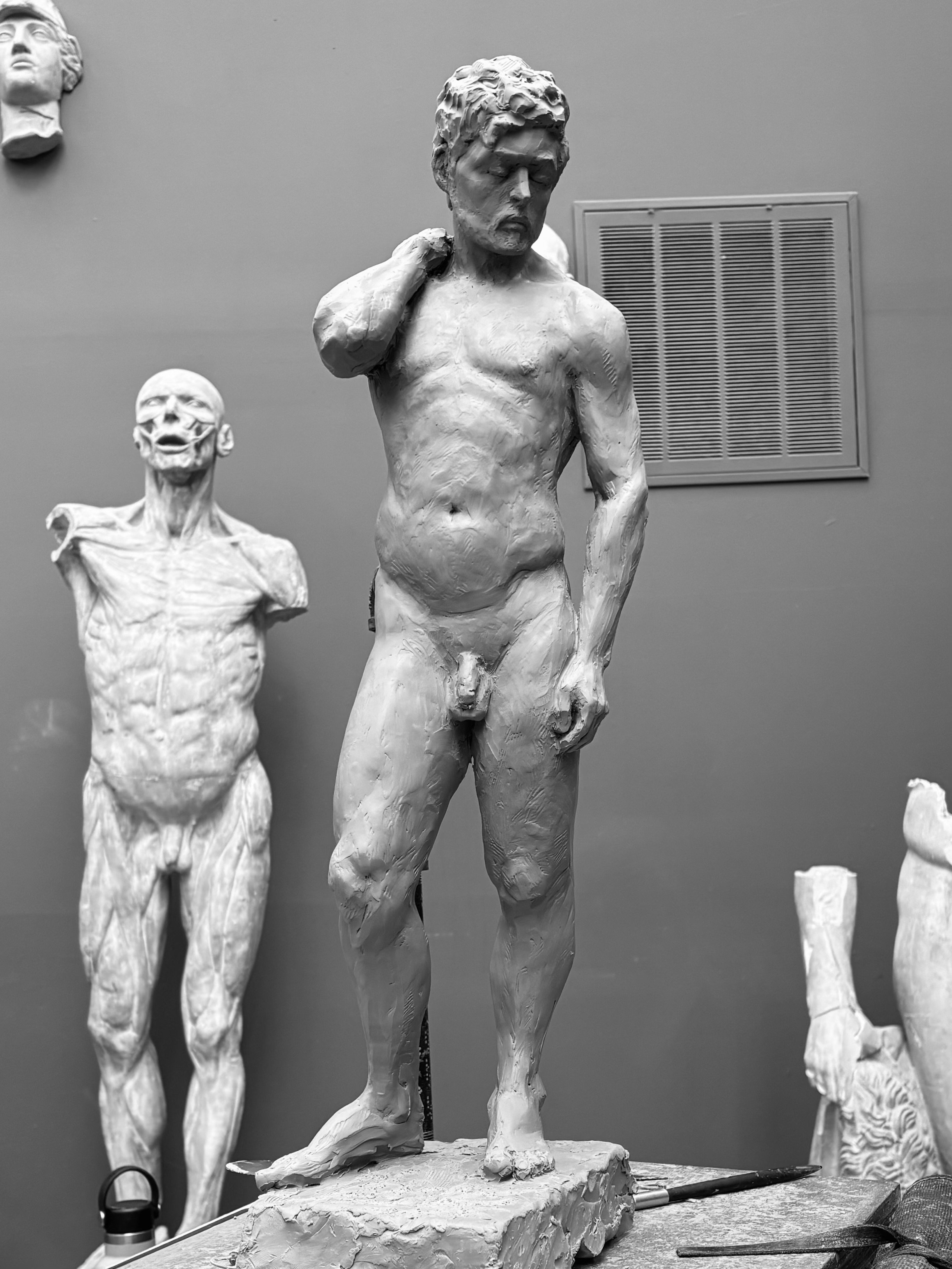 Black and white photo of a clay statue of a male figure with one hand behind his neck, surrounded by other sculptures.
