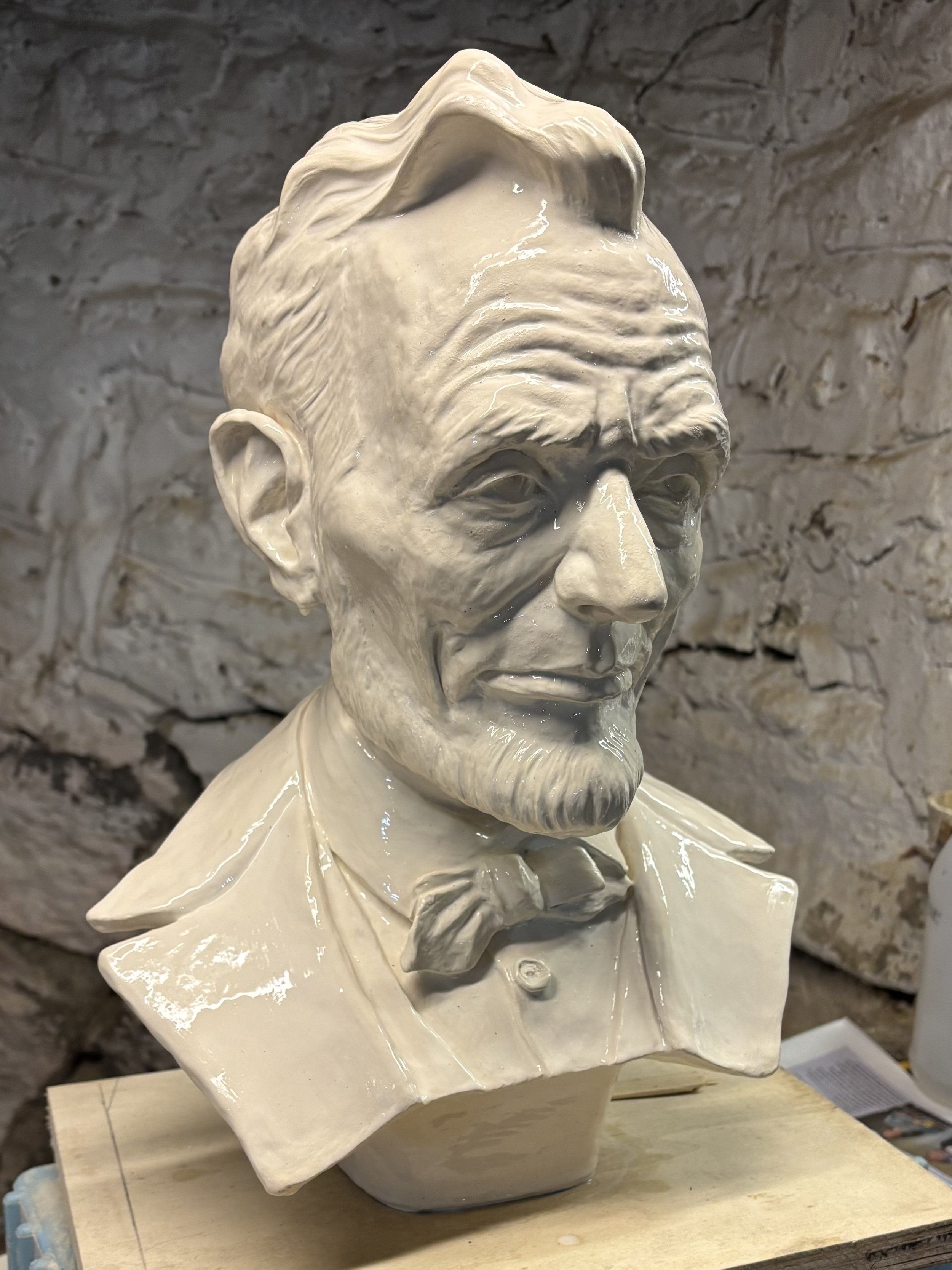 A white ceramic bust sculpture of Abraham Lincoln with a detailed face, hair, and clothing, displayed on a wooden surface against a stone wall background.