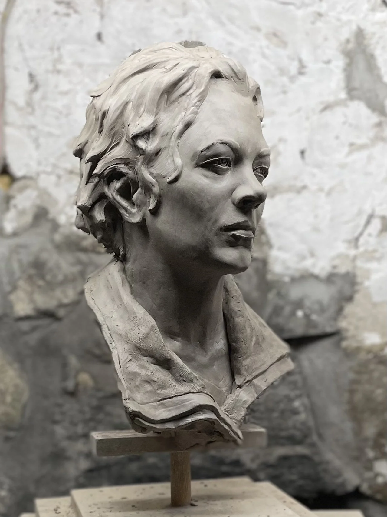 Clay bust sculpture of a woman with short, wavy hair, wearing a collared shirt, set against a stone wall background.