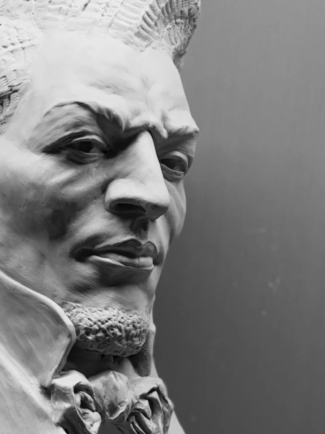 Close-up of a clay bust sculpture of historical figure Frederick Douglass with a beard and mustache, wearing a coat and bow tie, against a plain background.