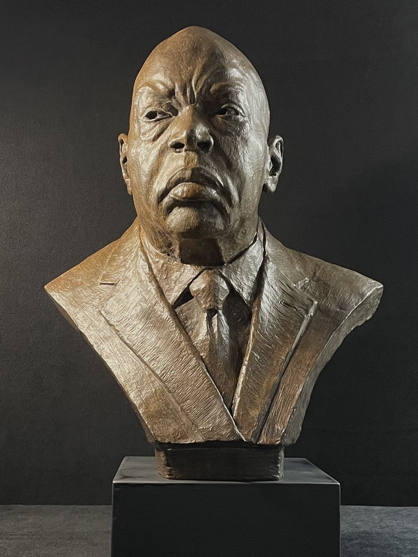 Bronze bust of a man with a serious expression, dressed in a suit and tie, set against a black background.