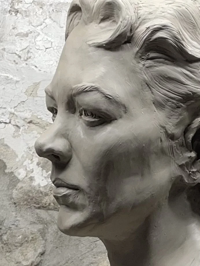 Close-up of a gray clay sculpture of a woman's face in profile, with detailed features and textured hair, against a rough, white stone background.