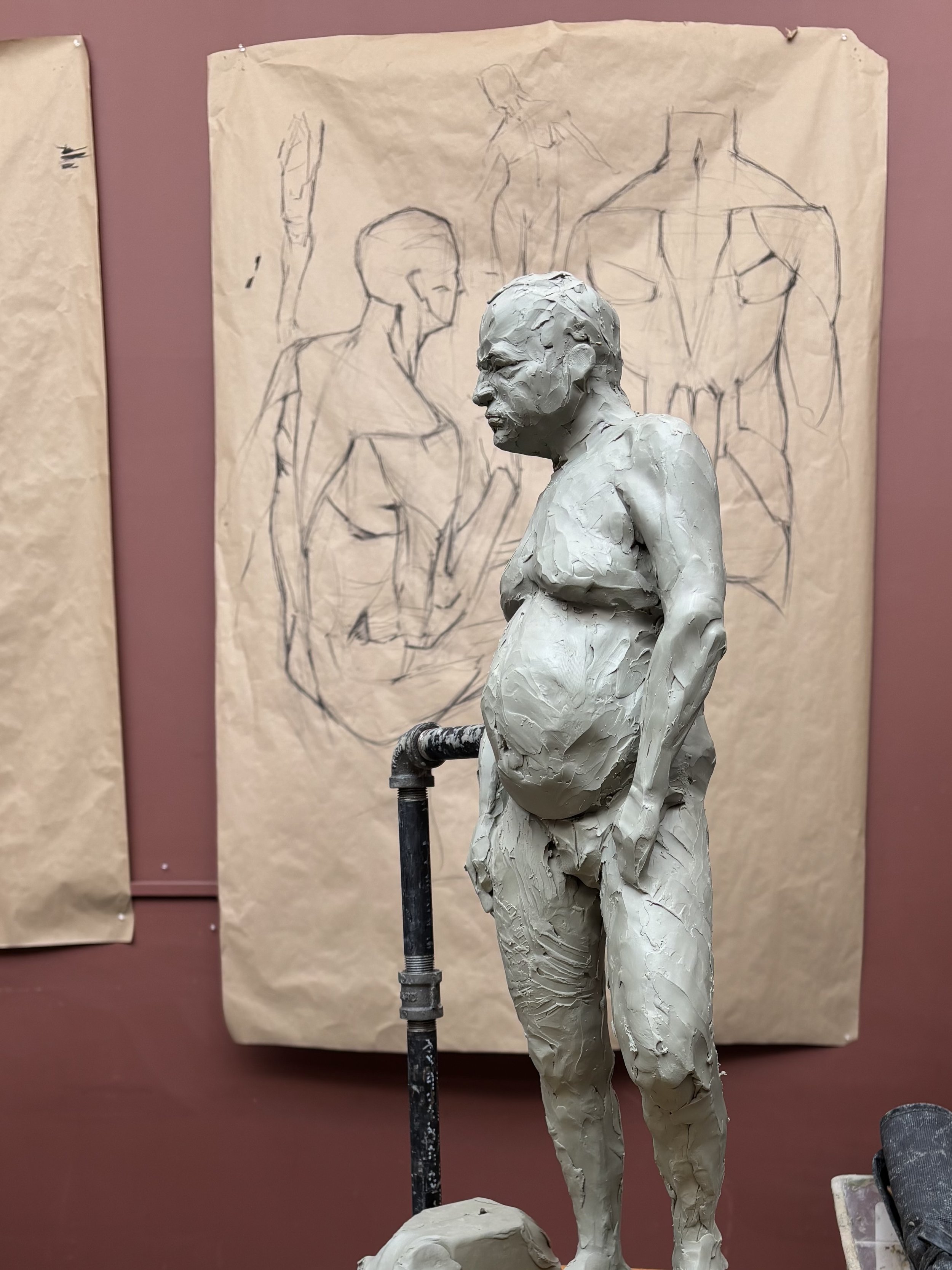 A sculpture of a man, crafted from clay. The background features sketches of various human figures on brown paper.