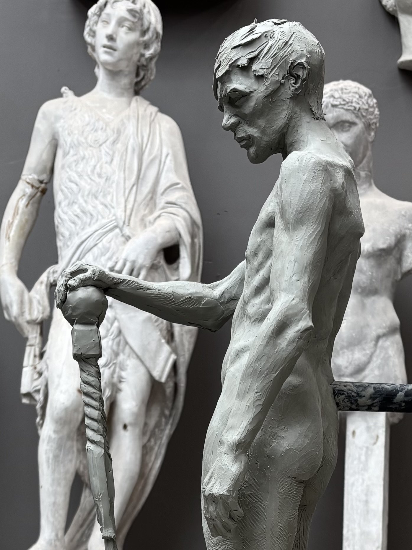 Several detailed white sculptures of nude male and female figures are displayed against a dark background, with the focus on one seated male sculpture with textured hair and facial features, and a standing female sculpture with short hair and a flowi