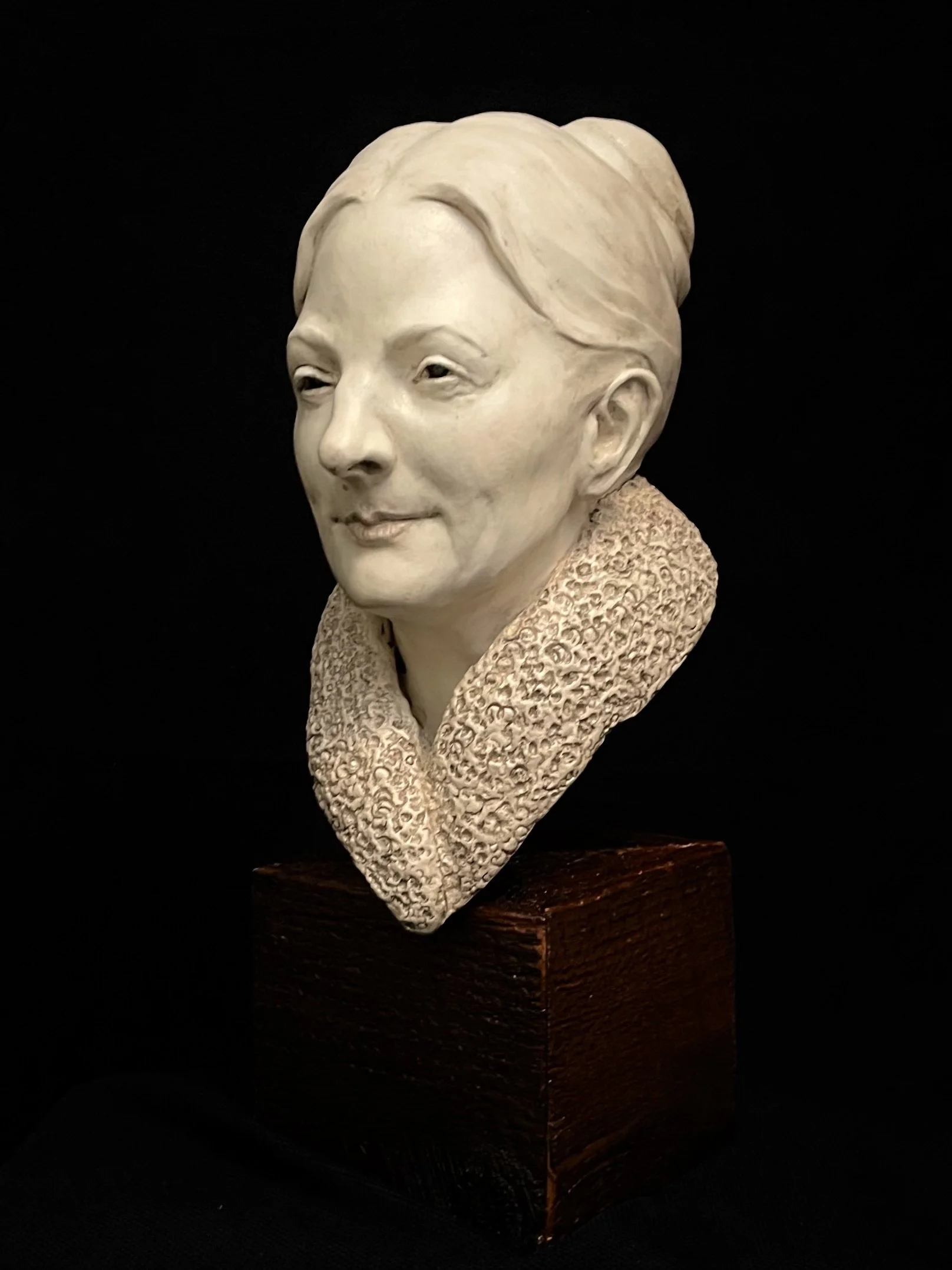 A white bust of a woman with short hair on a wooden base, against a black background.