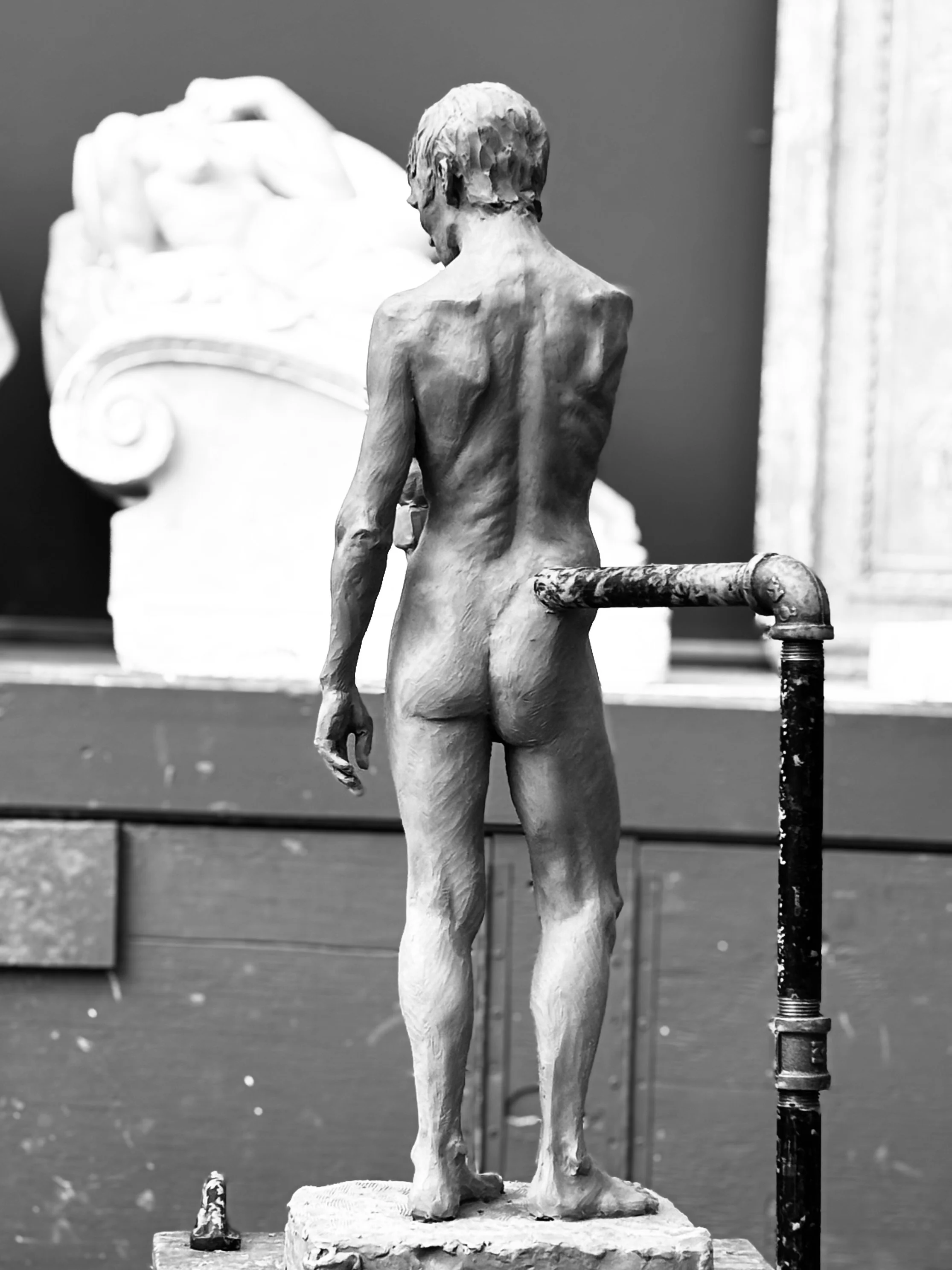 A black and white photo of a classical sculpture of a nude male figure viewed from the back, standing on a stone base, with an art gallery or museum setting in the background.