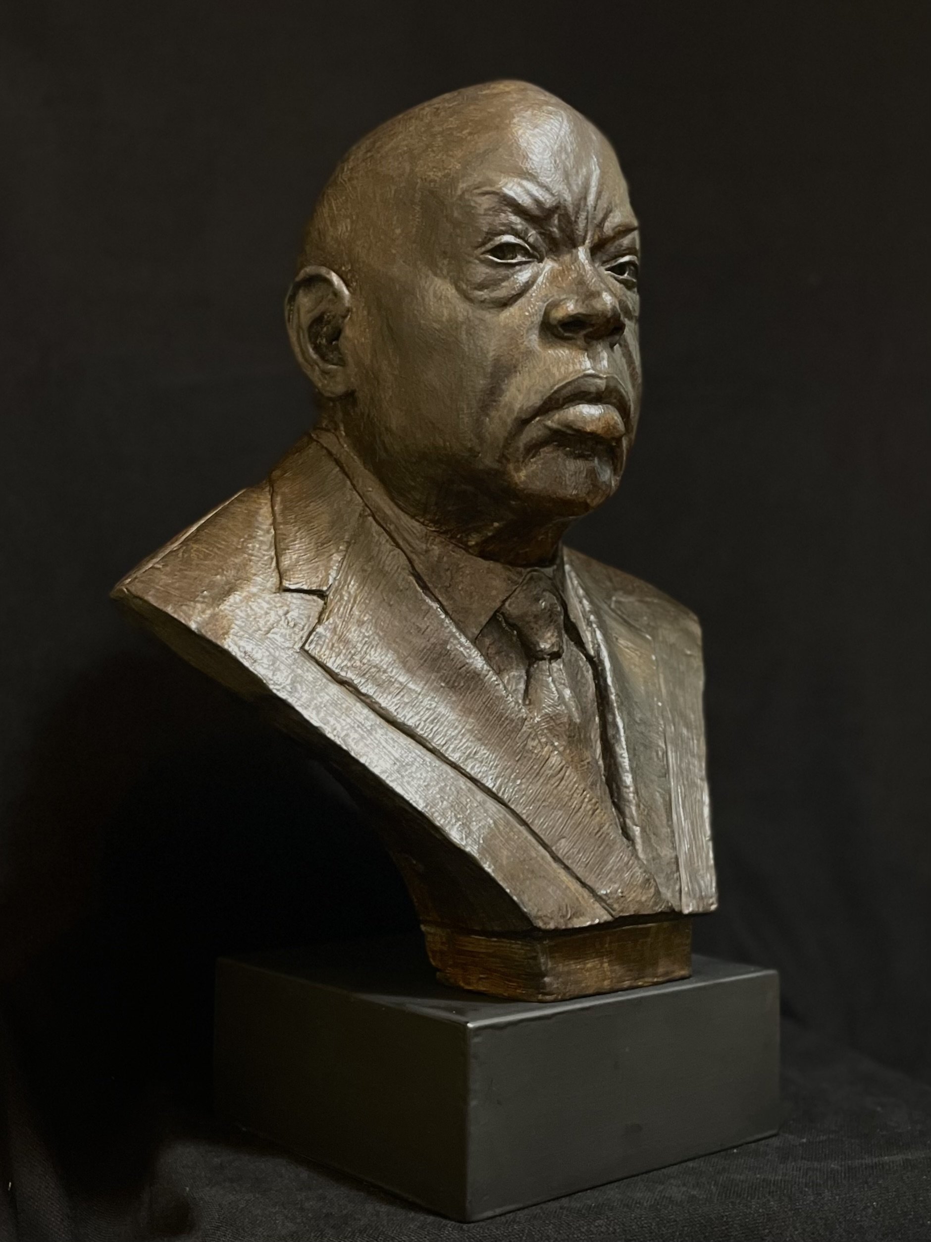 Bronze bust sculpture of John Lewis, a man with a stern facial expression, dressed in a suit, placed on a black pedestal against a dark background.