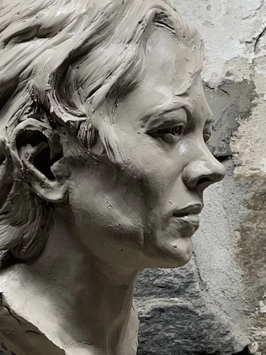Close-up of a sculpted bust of a woman in profile, showing detailed facial features and textured hair, against a rough stone background.