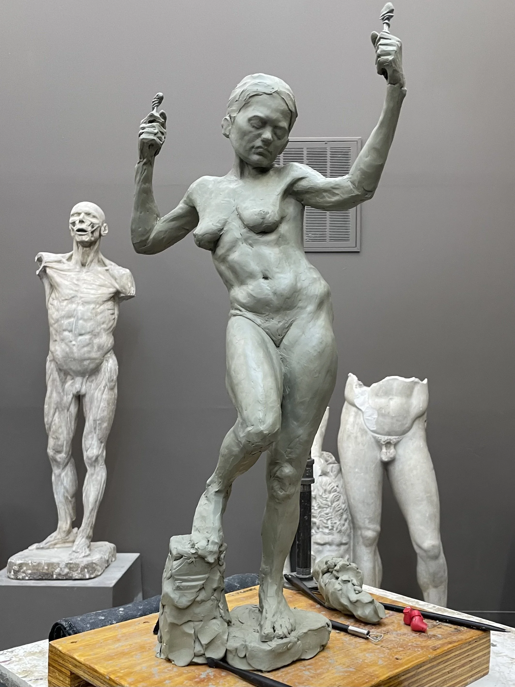 Clay sculpture of a woman with arms raised, surrounded by other clay sculptures of men and women in various poses, on a wooden table in an art studio.