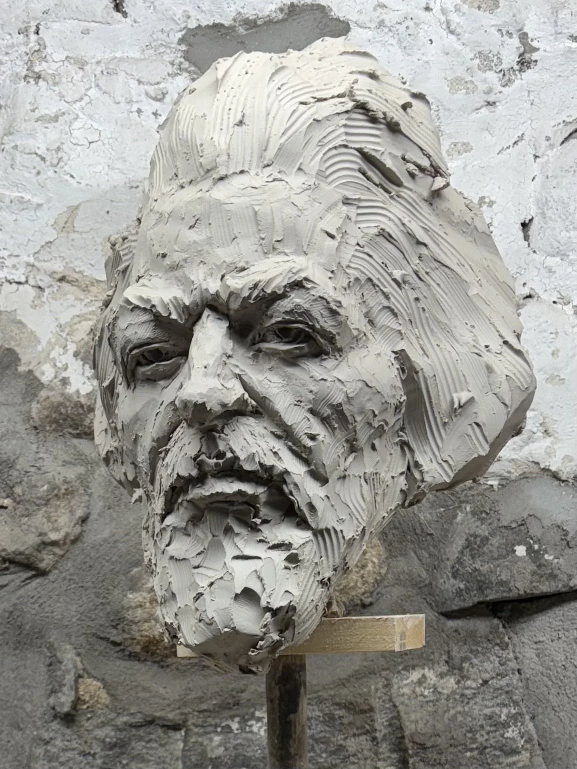 A sculpture of a human face sculpted from clay, with detailed features and textured hair and beard, set against a rough stone background.
