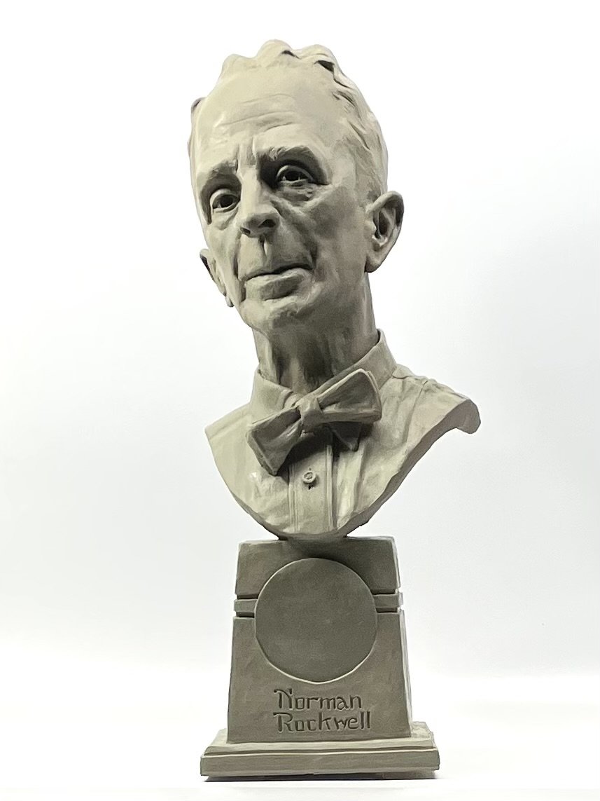A white sculpted bust of Norman Rockwell with a serious expression, wearing a collared shirt and bow tie, mounted on a pedestal with his name engraved on it.