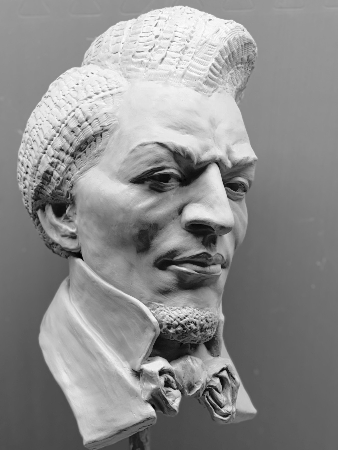 Black and white sculpture bust of Frederick Douglass, a man with styled hair, mustache, beard, and formal attire, against a plain background.