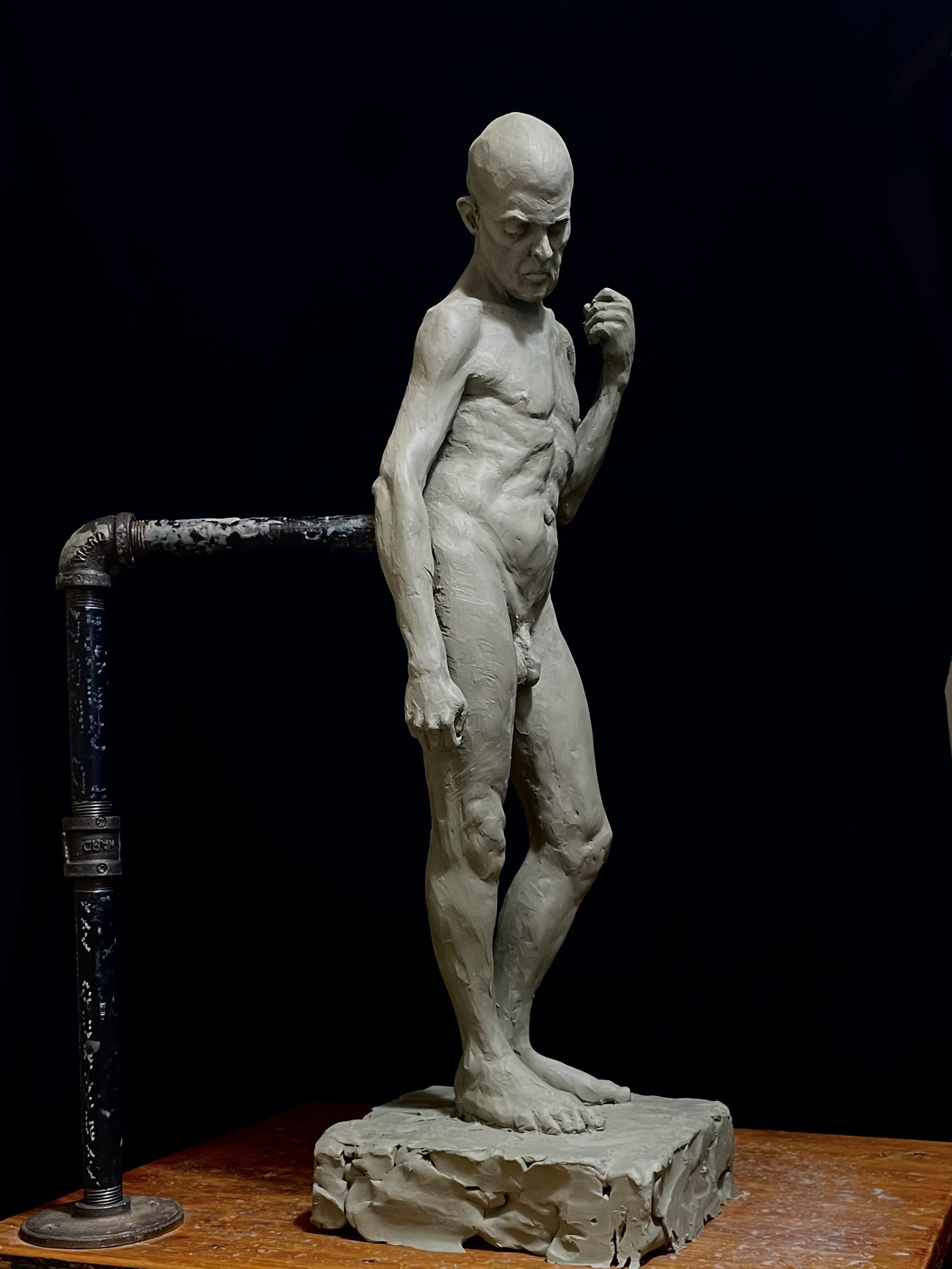A sculpture of a man with a bald head, muscular body, and clenched fist, standing on a rough stone base, in front of a dark background.