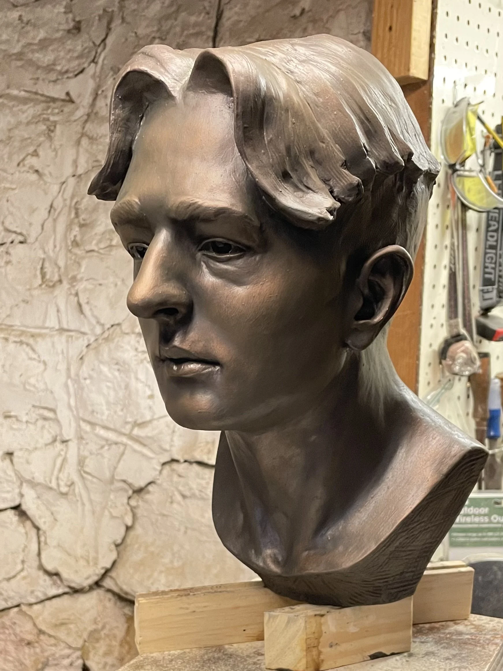Bronze bust sculpture of a young man with short hair, mounted on a wooden base, in a workshop or gallery with a stone wall background.