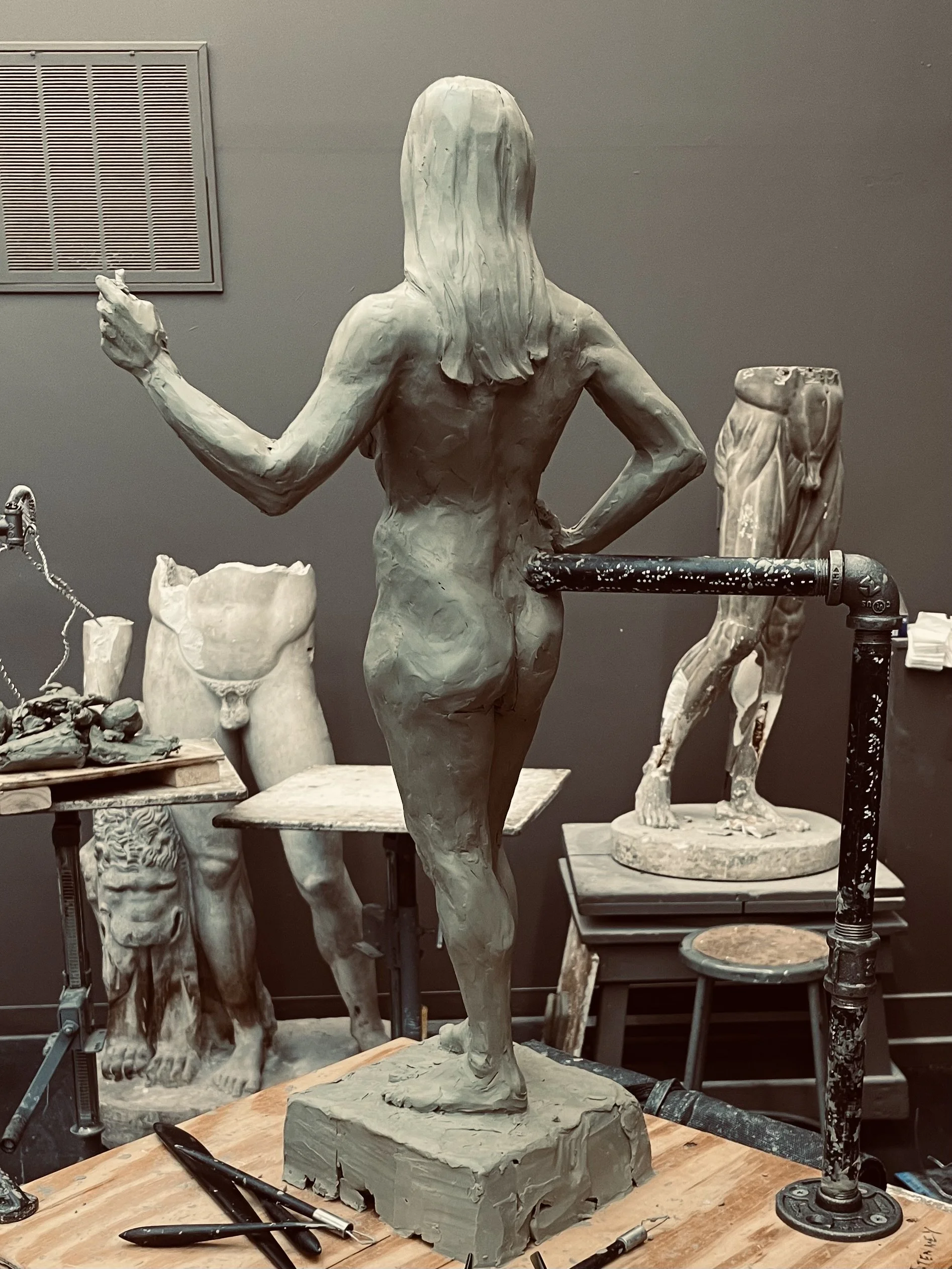 A sculpture of a woman with one arm raised and the other on her hip, in an artist's studio surrounded by other sculpture pieces and tools.