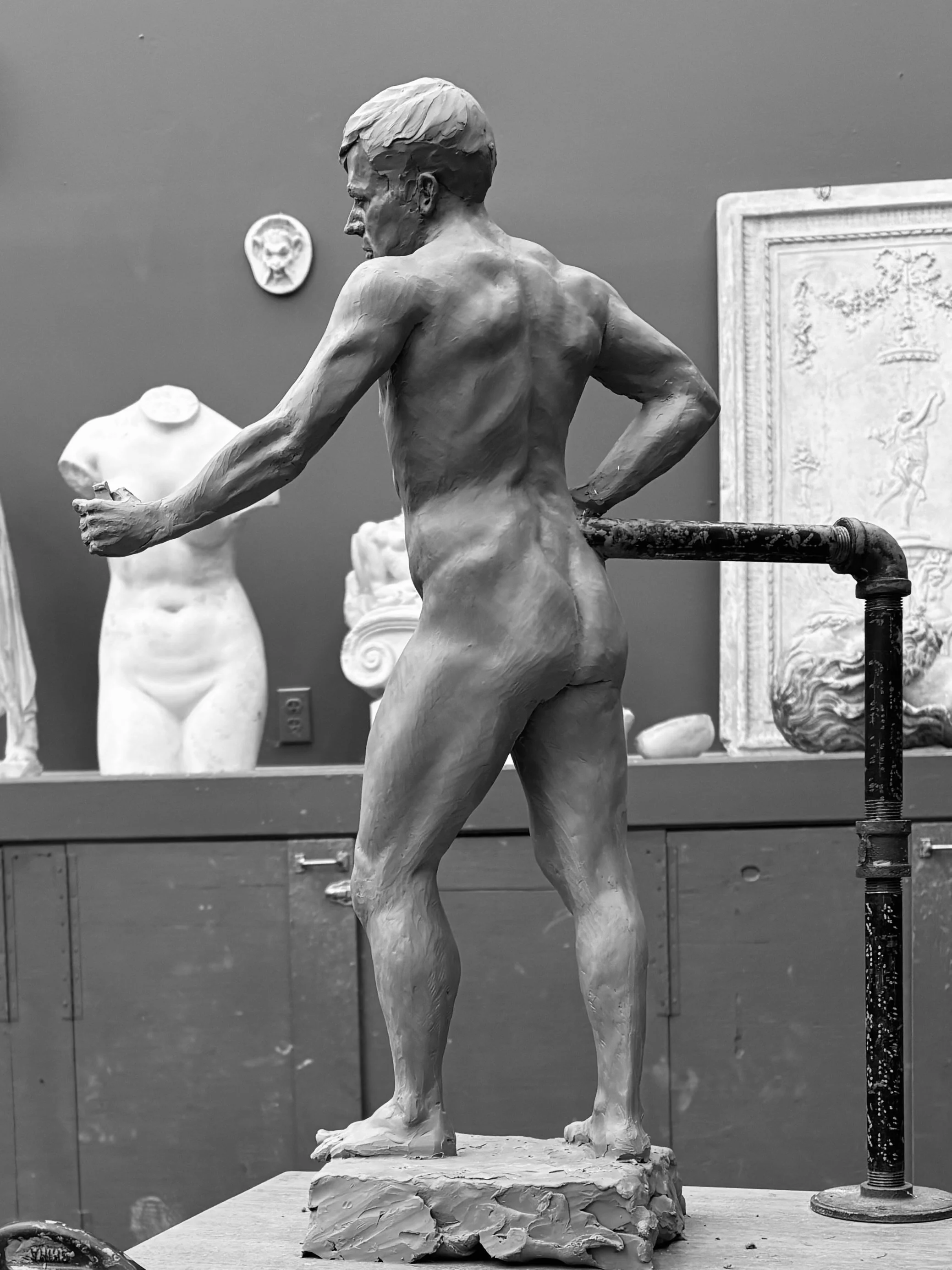 Black and white photo of a classical-style sculpture of a male figure, viewed from behind, standing on a textured base in an art studio surrounded by other statues and art pieces.