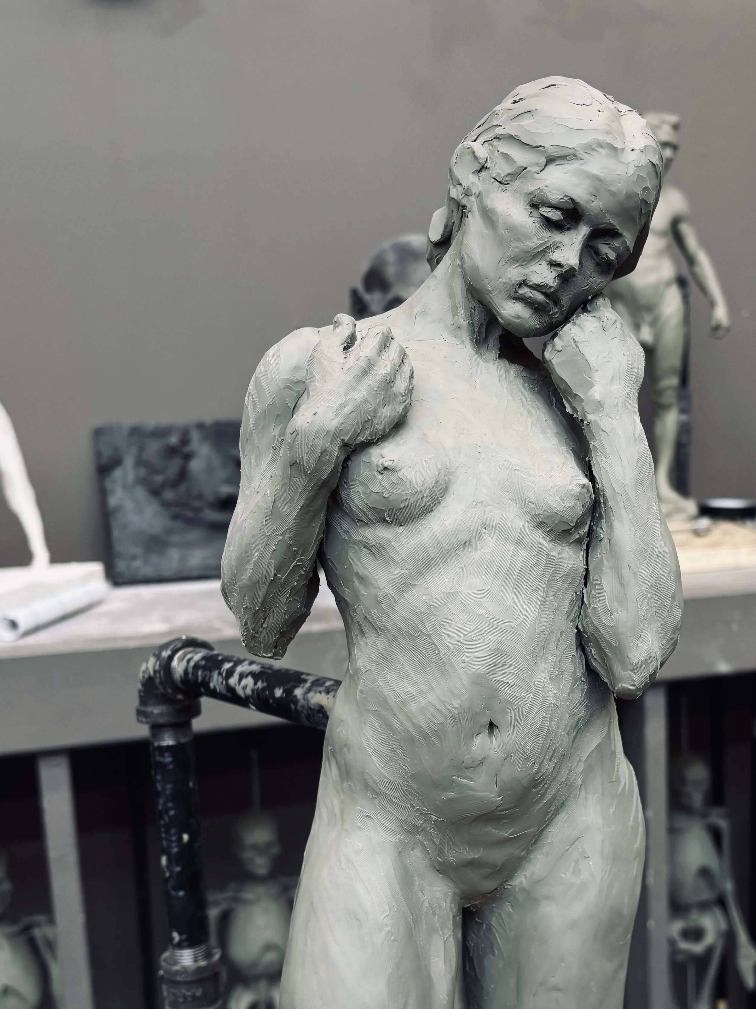 A sculpture of a woman with her hand on her shoulder, made with rough, textured clay, with other similar sculptures in the background.