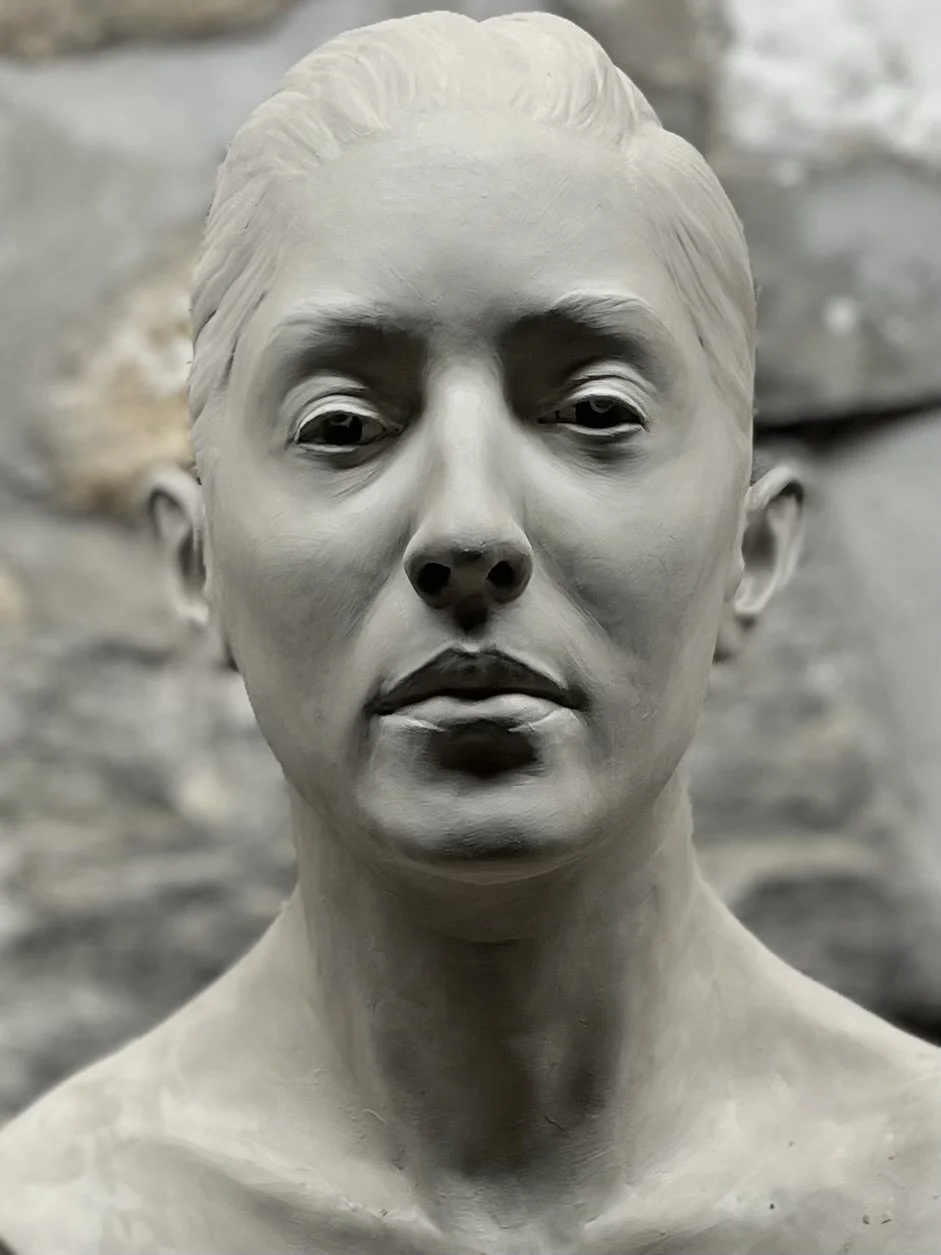 Clay sculpture of a woman's face with a neutral expression, detailed hair, and bare shoulders.