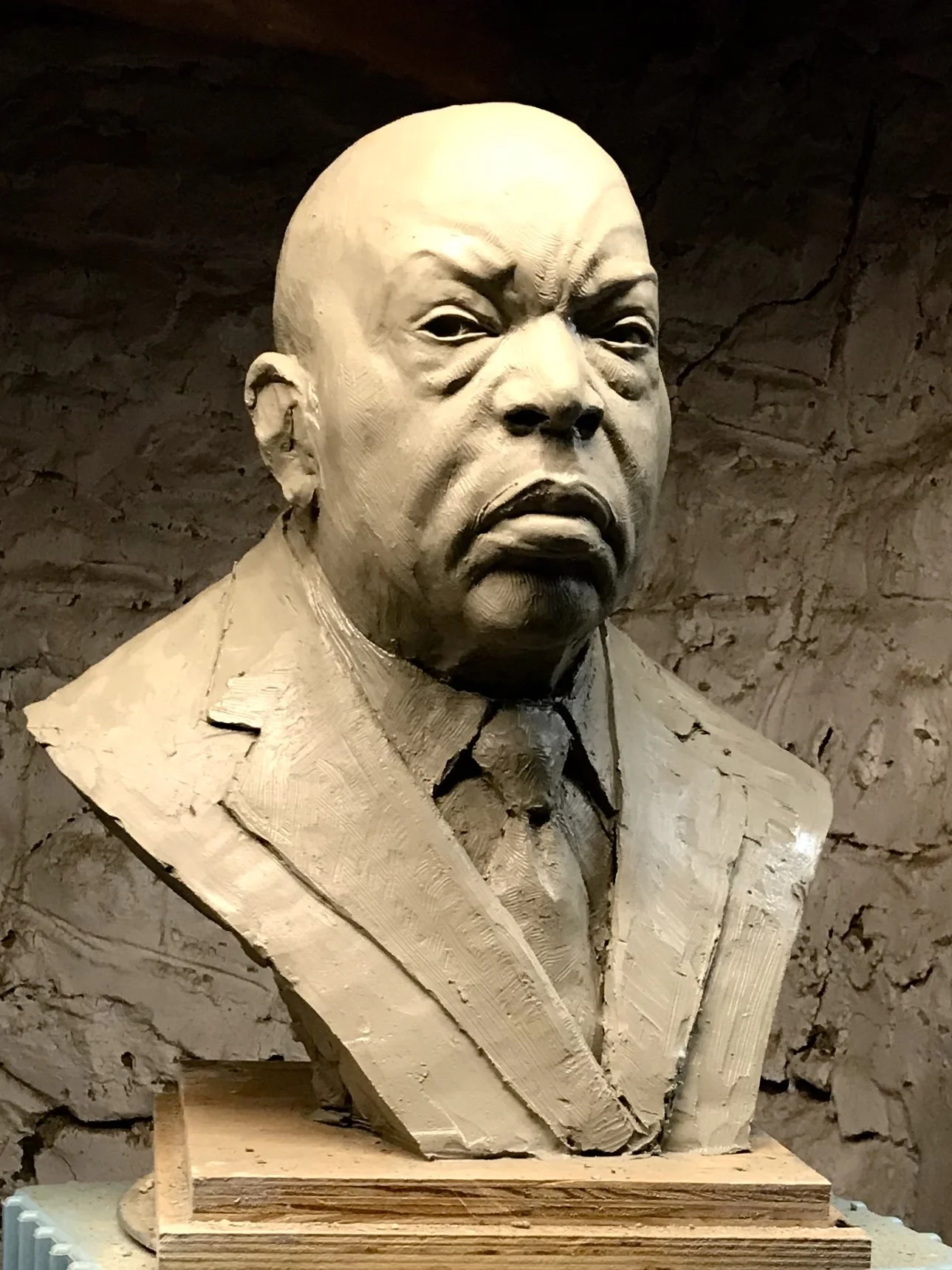 Clay bust sculpture of John Lewis, a stern-looking man in a suit, displayed against a textured stone wall.