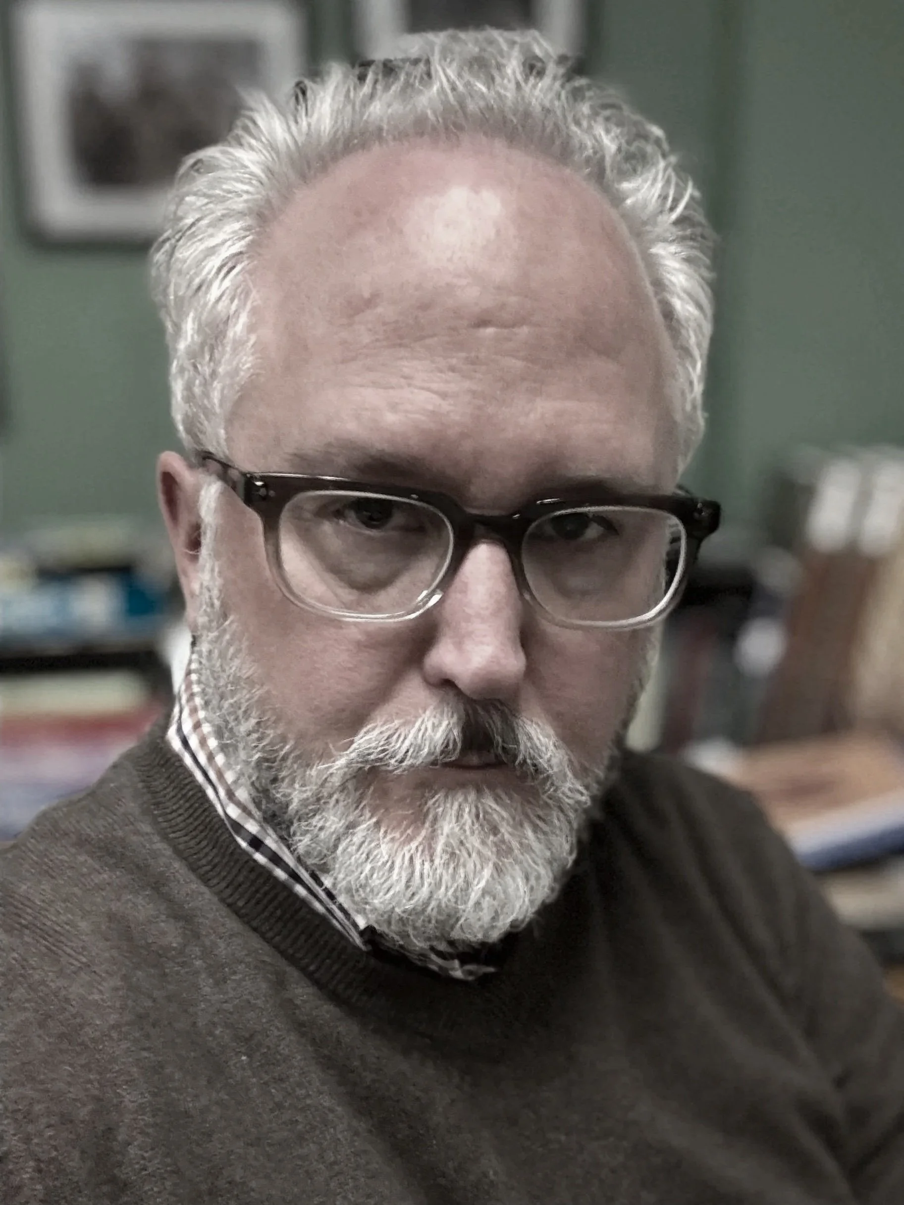 Close-up of an older man with a gray beard and glasses, wearing a checkered shirt and a brown sweater, in an indoor setting.
