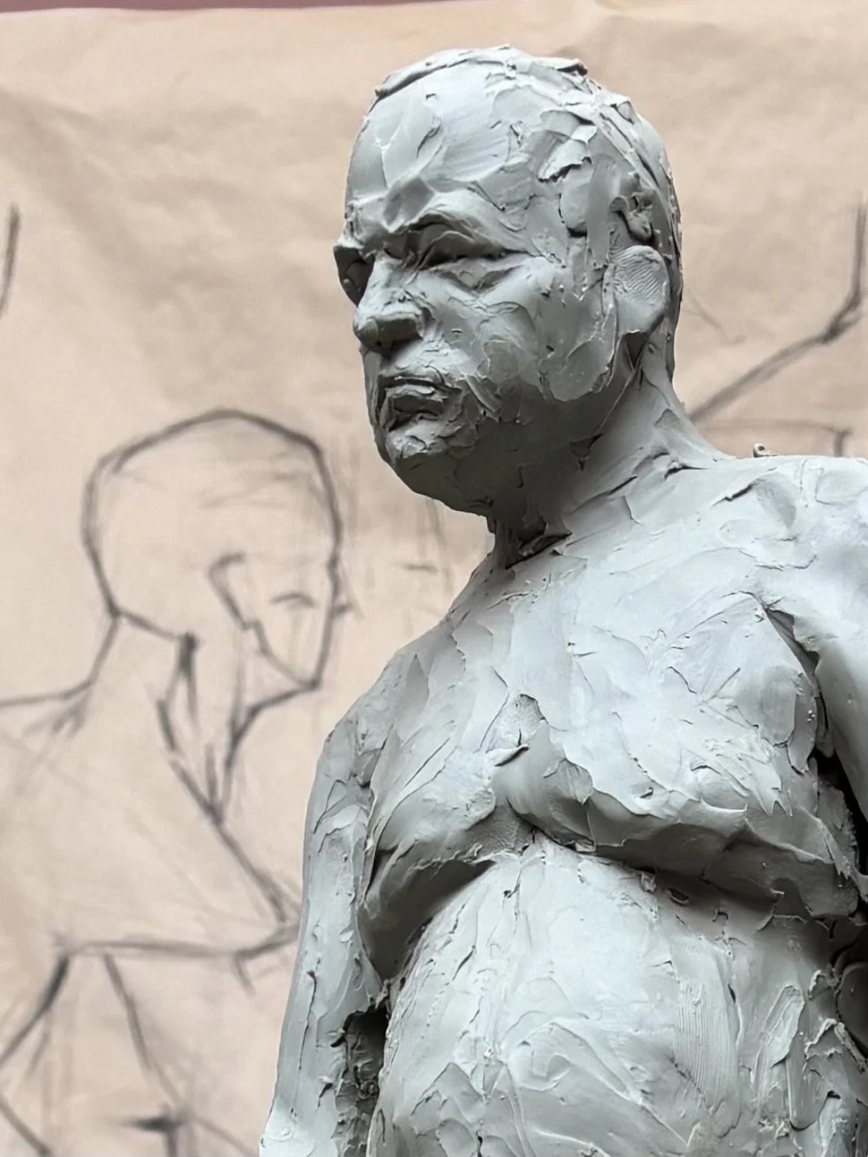 A clay sculpture of a person's upper body and face with a serious expression, situated in front of just a sketch of a head and shoulders in the background.