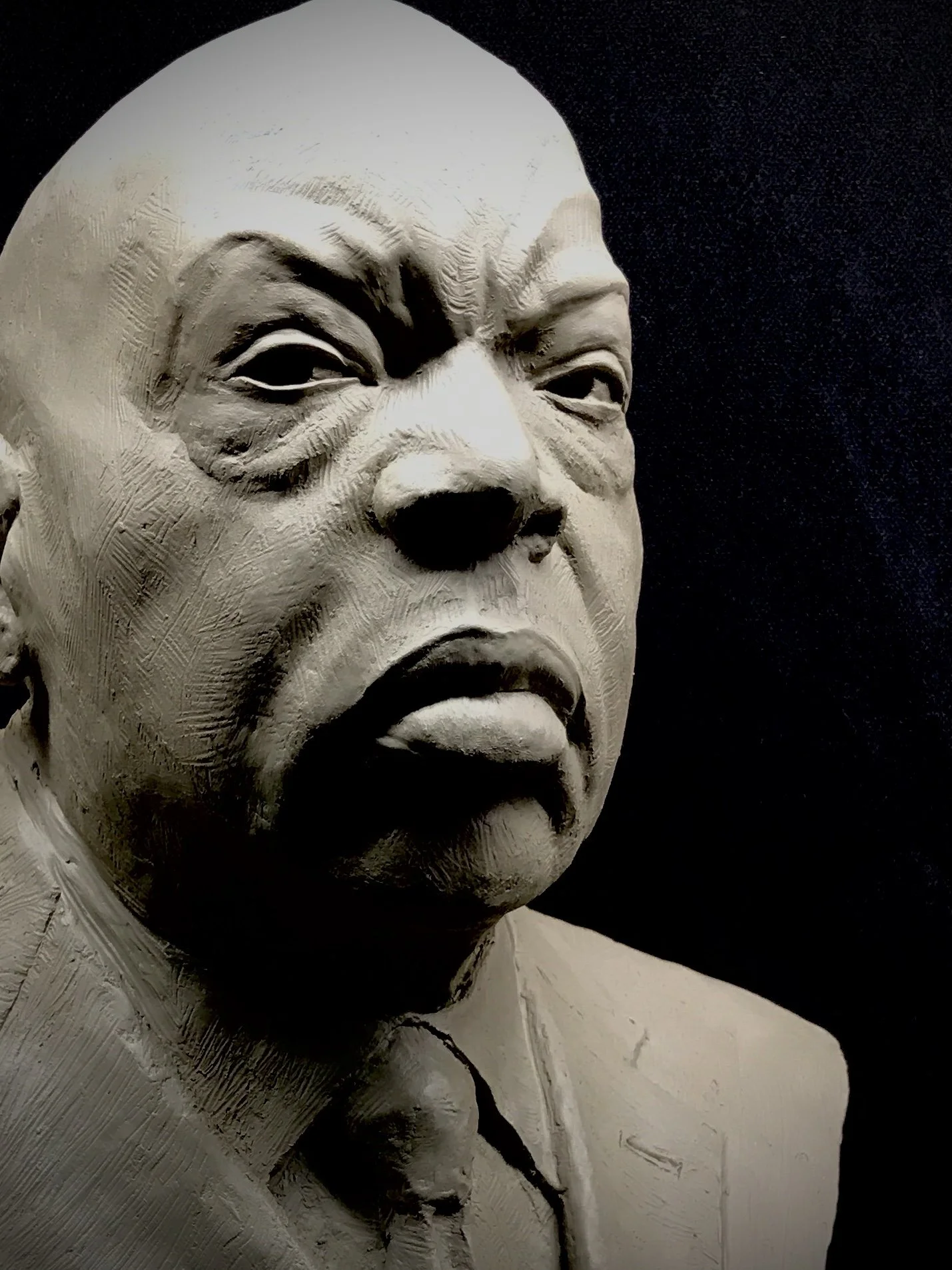 Close-up of a textured clay sculpture of John Lewis, showing his face and upper body against a dark background.