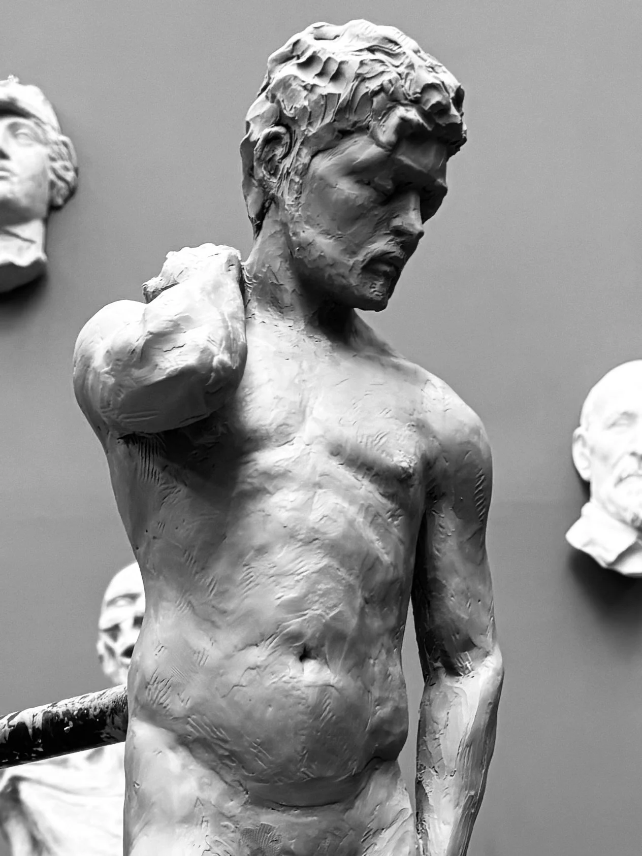 A sculpture of a male torso with a detailed face, hair, and muscular build, shown in profile against a gray background, with other statue busts partially visible in the background.