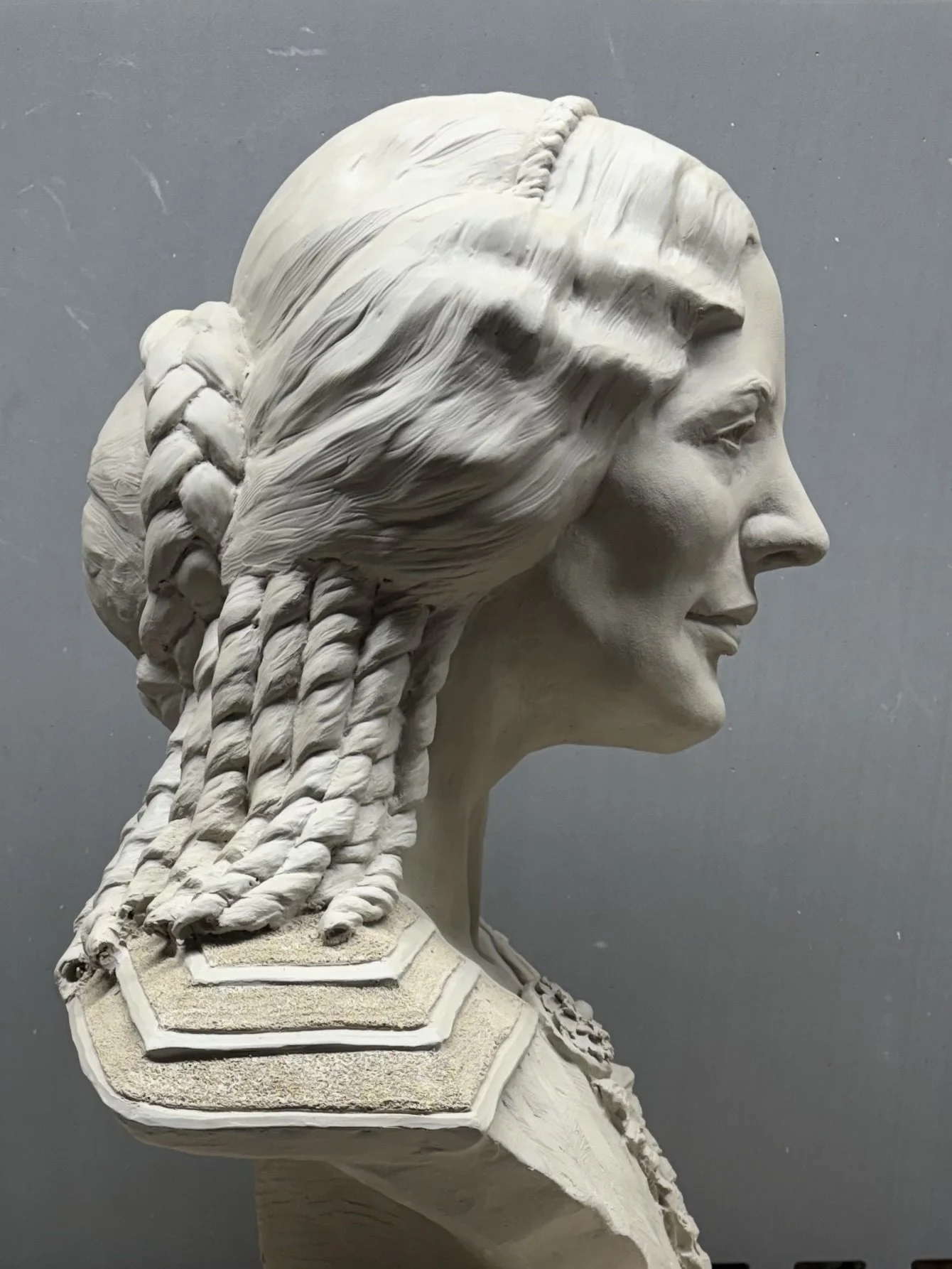A clay sculpted bust of a woman with braided hair, facing right, against a gray background.
