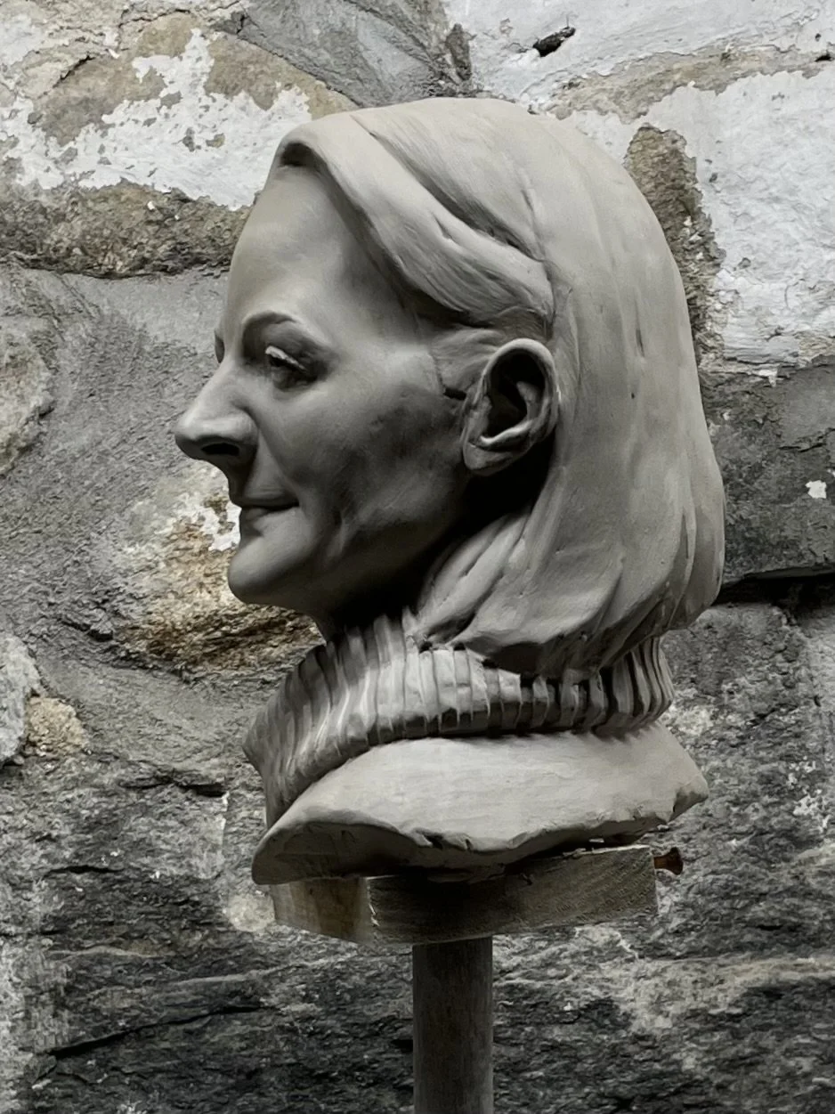 A detailed sculpture of a woman's head and shoulders, with slicked-back hair and a high-collared top, mounted on a stand against a rough stone wall background.