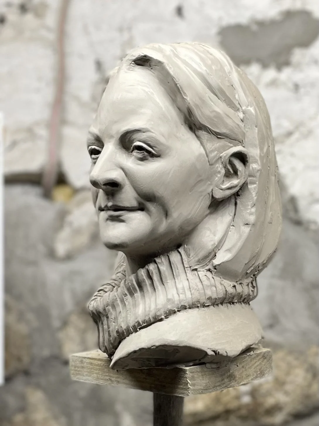Unfinished clay sculpture of a woman's bust with detailed facial features and hair, mounted on a wooden stand, set against a brick wall background.