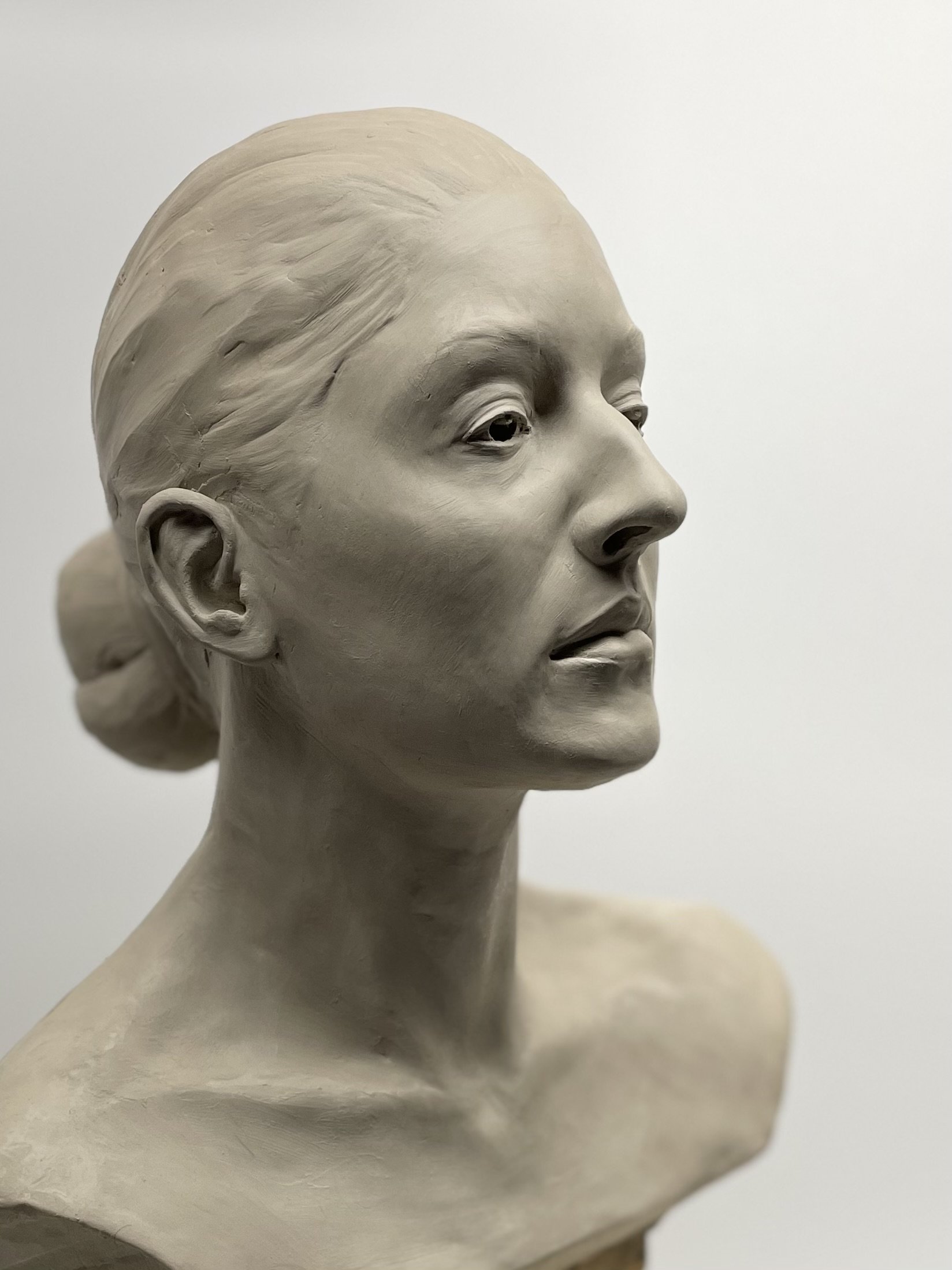 Close-up of a sculpture of a woman's face and upper shoulders with smooth features and detailed hair, set against a plain background.