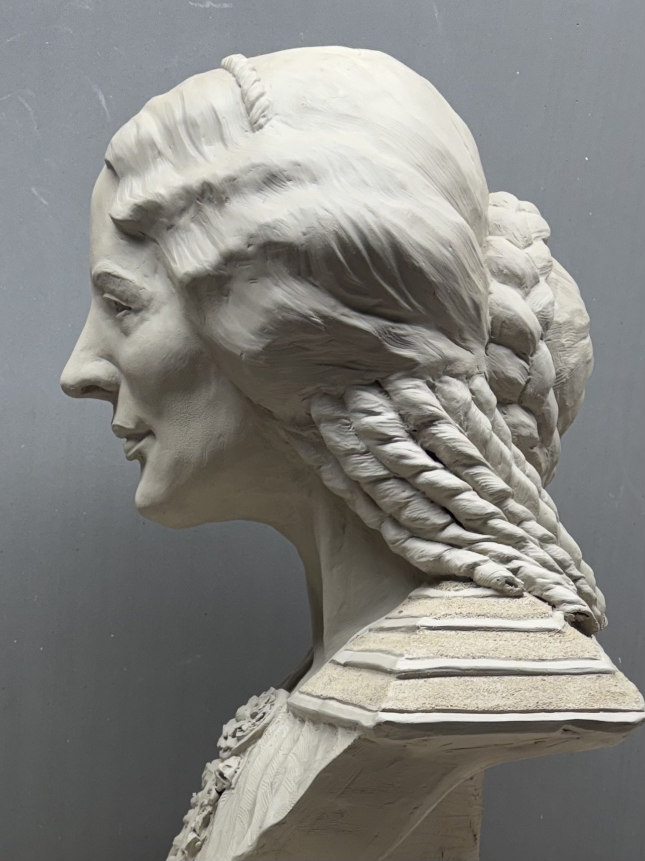 Side profile of a clay classical sculpture of a woman with braided hair, set against a gray background.