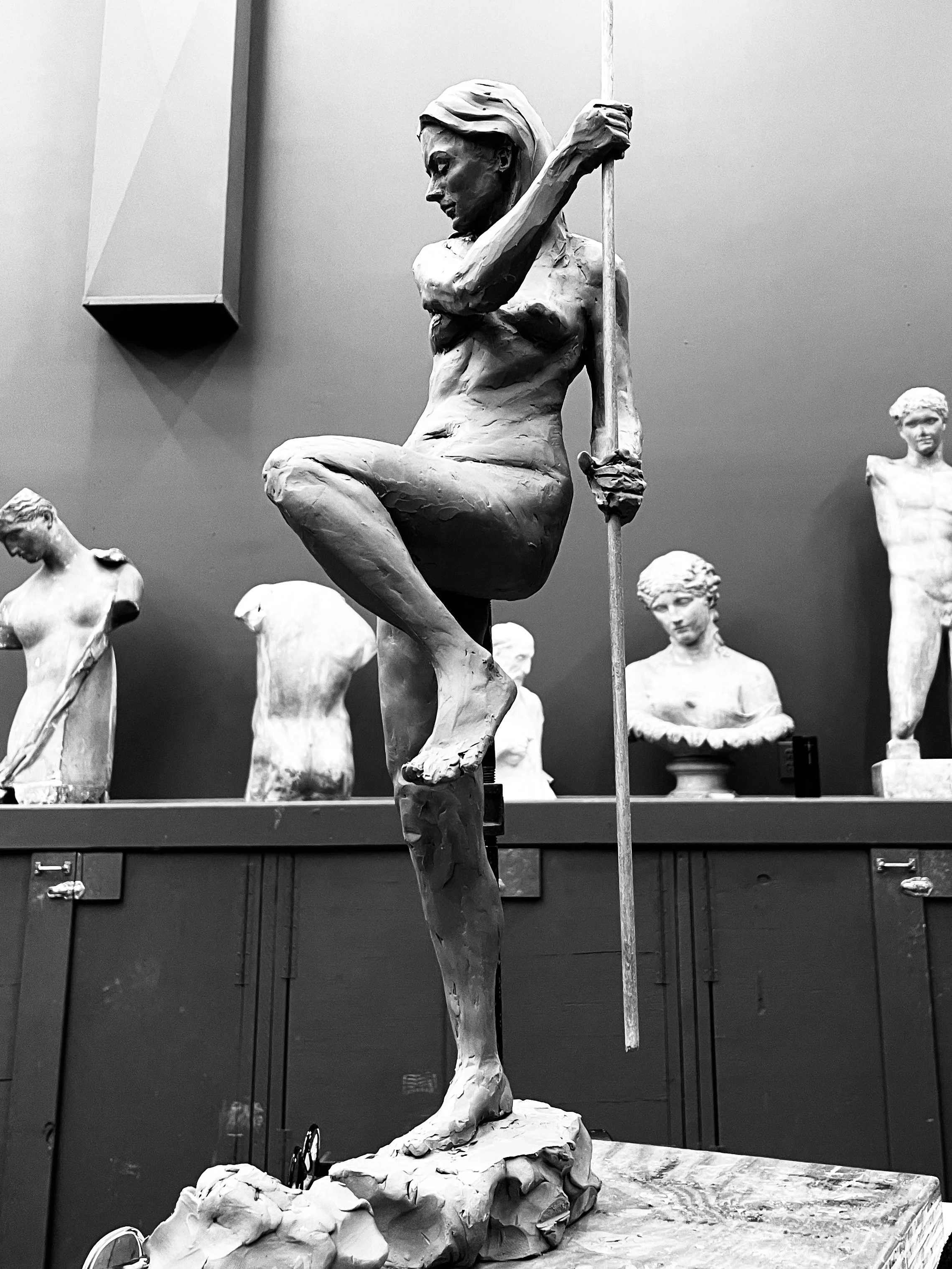 A sculpture of a woman holding a long staff, sitting with one arm resting on her knee, surrounded by several other sculptures of women in a gallery or museum setting.