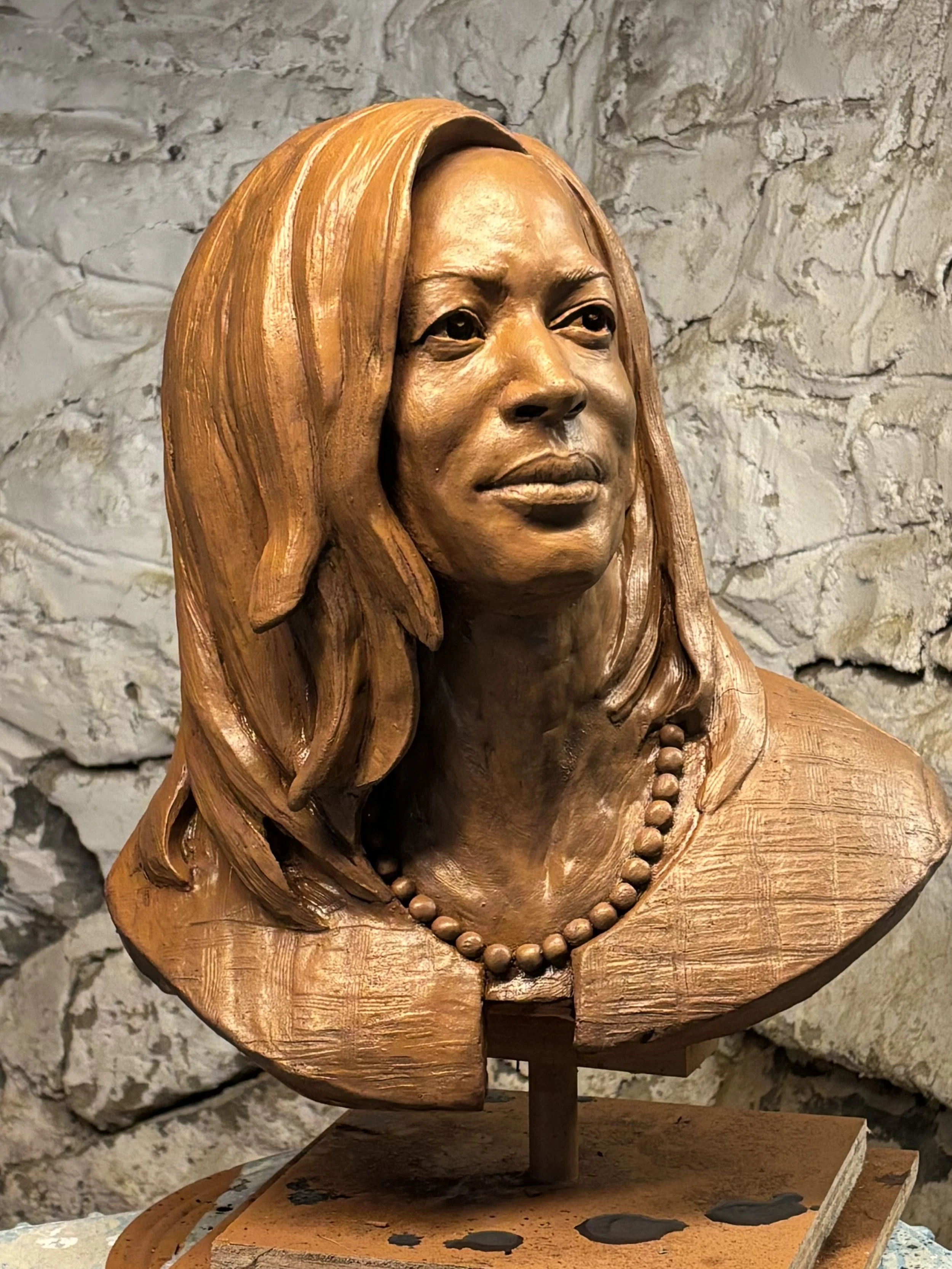 Wooden sculpture of a woman with long hair and a beaded necklace, displayed against a stone wall.