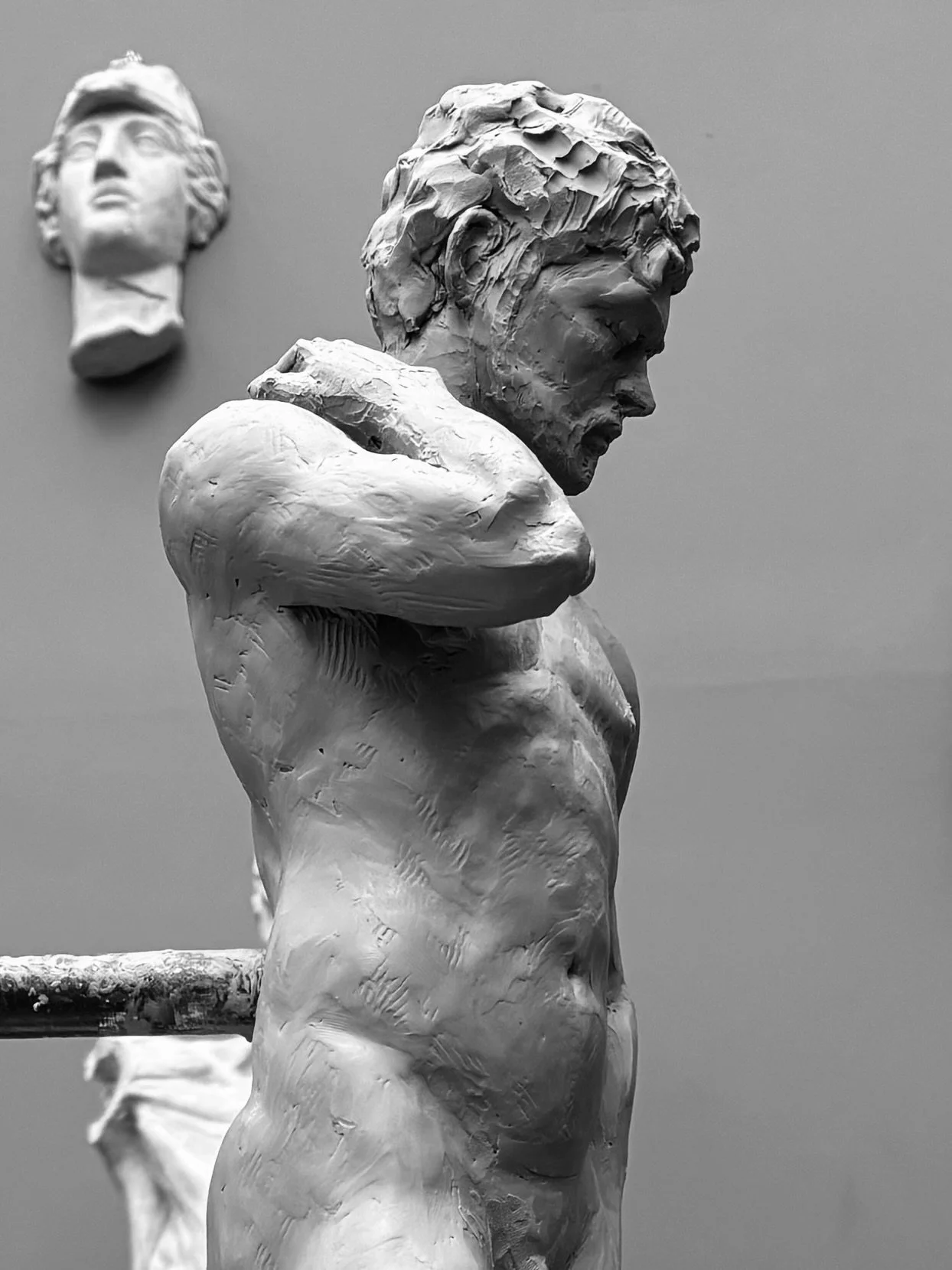A sculpture of a person in profile resting their chin on their crossed arms, with a second, smaller sculpture of a face in the background on the wall.