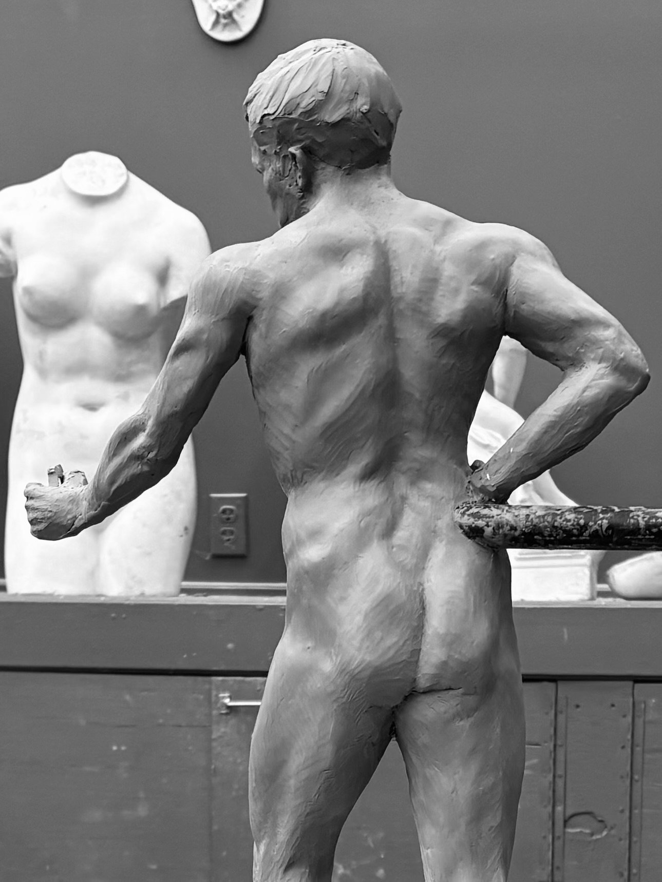Back view of a male sculpture in progress, with a female sculpture in the background in an art studio.