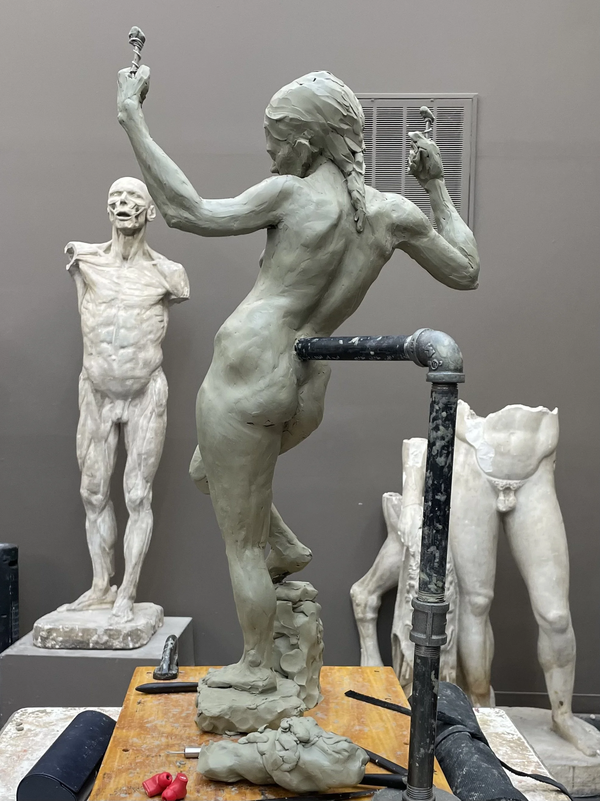 A sculpture of a muscular woman with long hair in a flexing pose, looking down and to the side, with a sculpture of a skeletal man in the background. The sculptures are in a workshop with tools and work materials on the table.