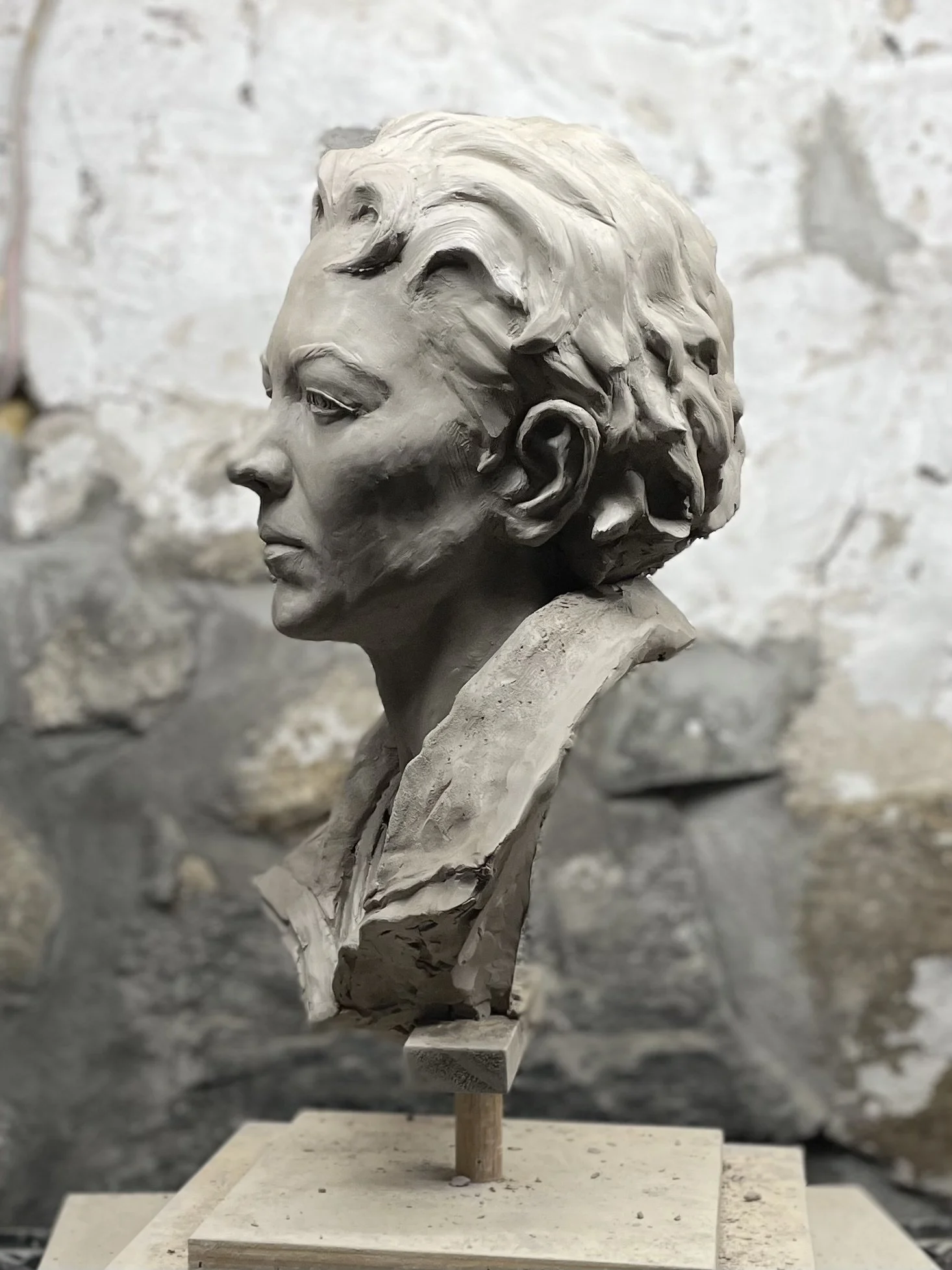 Clay sculpture of a woman's profile with detailed hair and facial features on a wooden stand against a textured background.