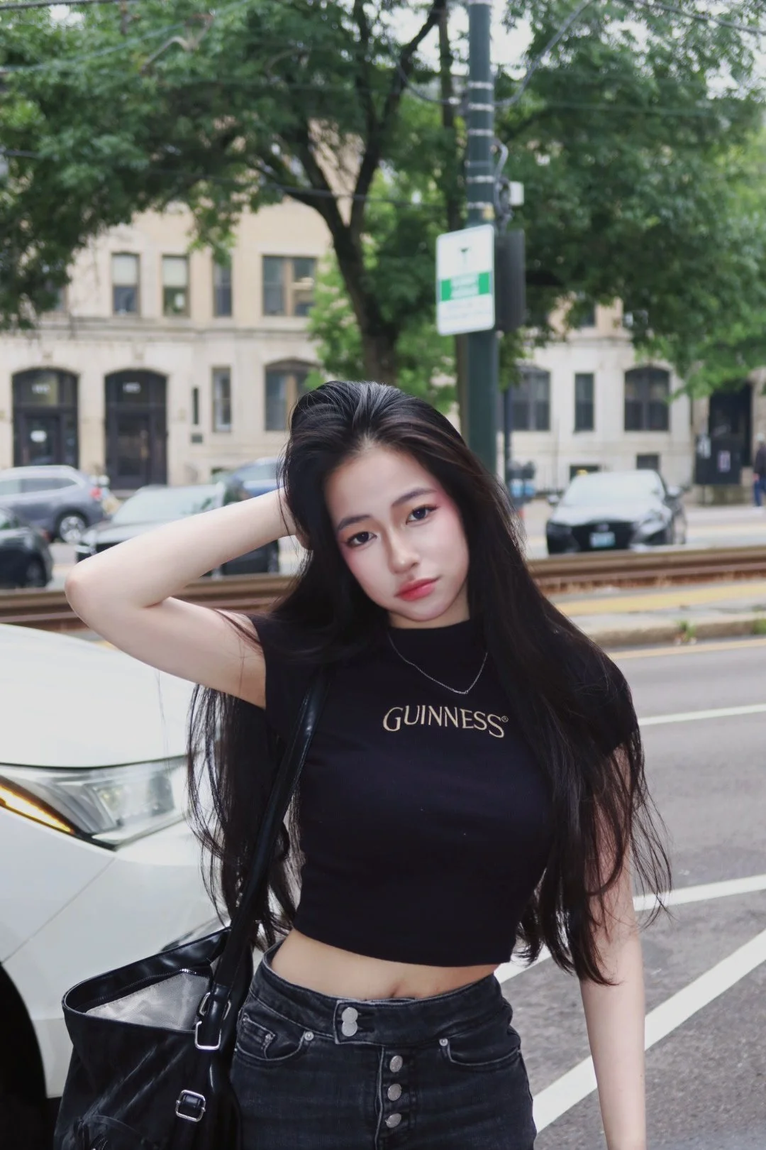 Young woman with long dark hair wearing a black "Guinness" crop top and black jeans posing outdoors near a white car, with trees and buildings in the background.