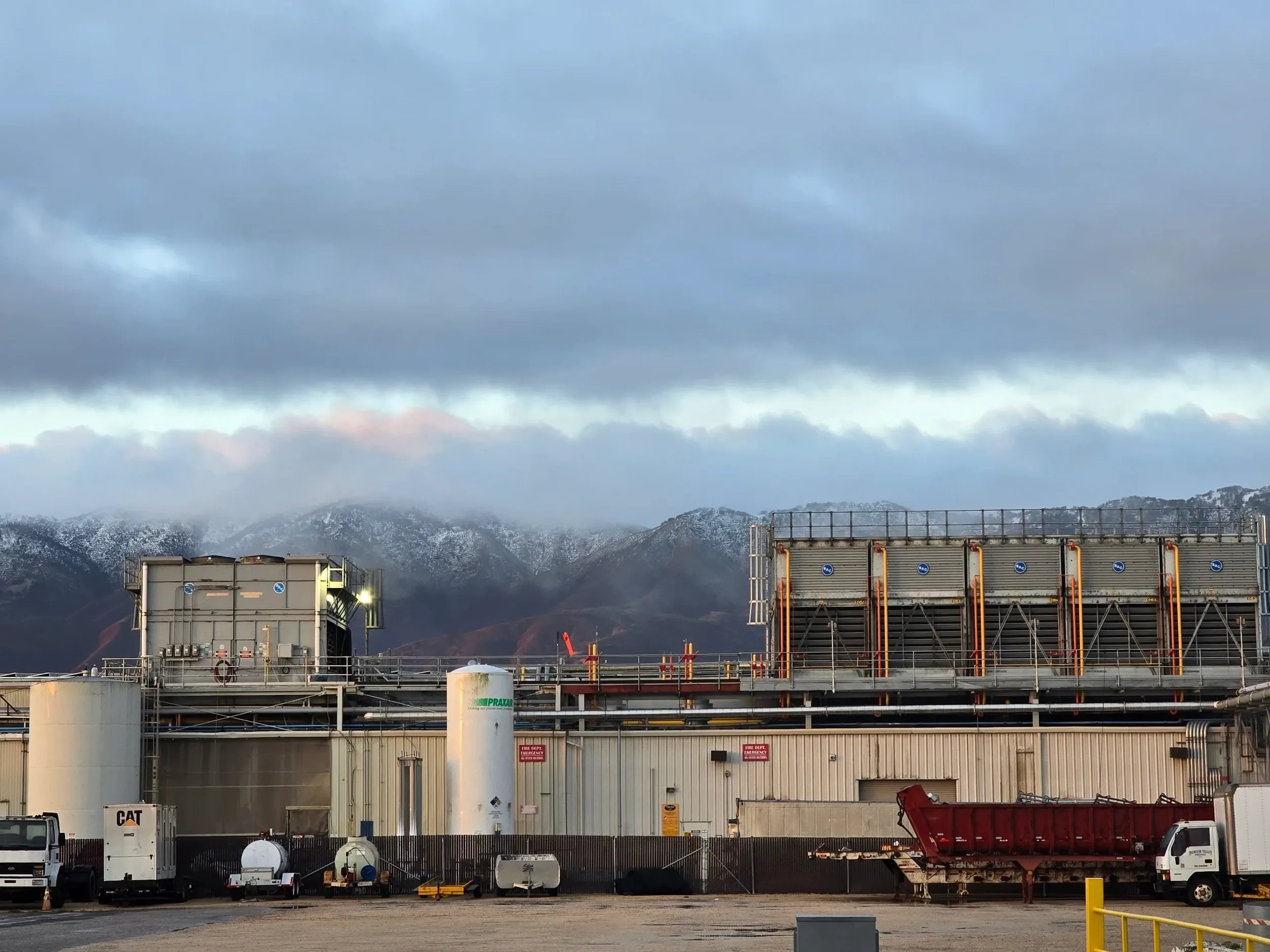 Industrial Facility with warehouses, storage tanks, and trucks, set against a mountainous background under a cloudy sky.
