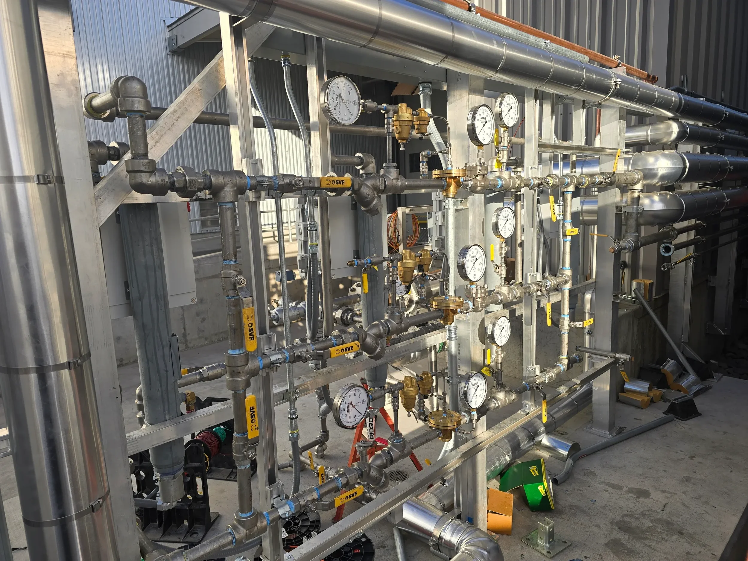 Close-up of industrial piping system with gauges, valves, and fittings mounted on a metal framework outside a building.