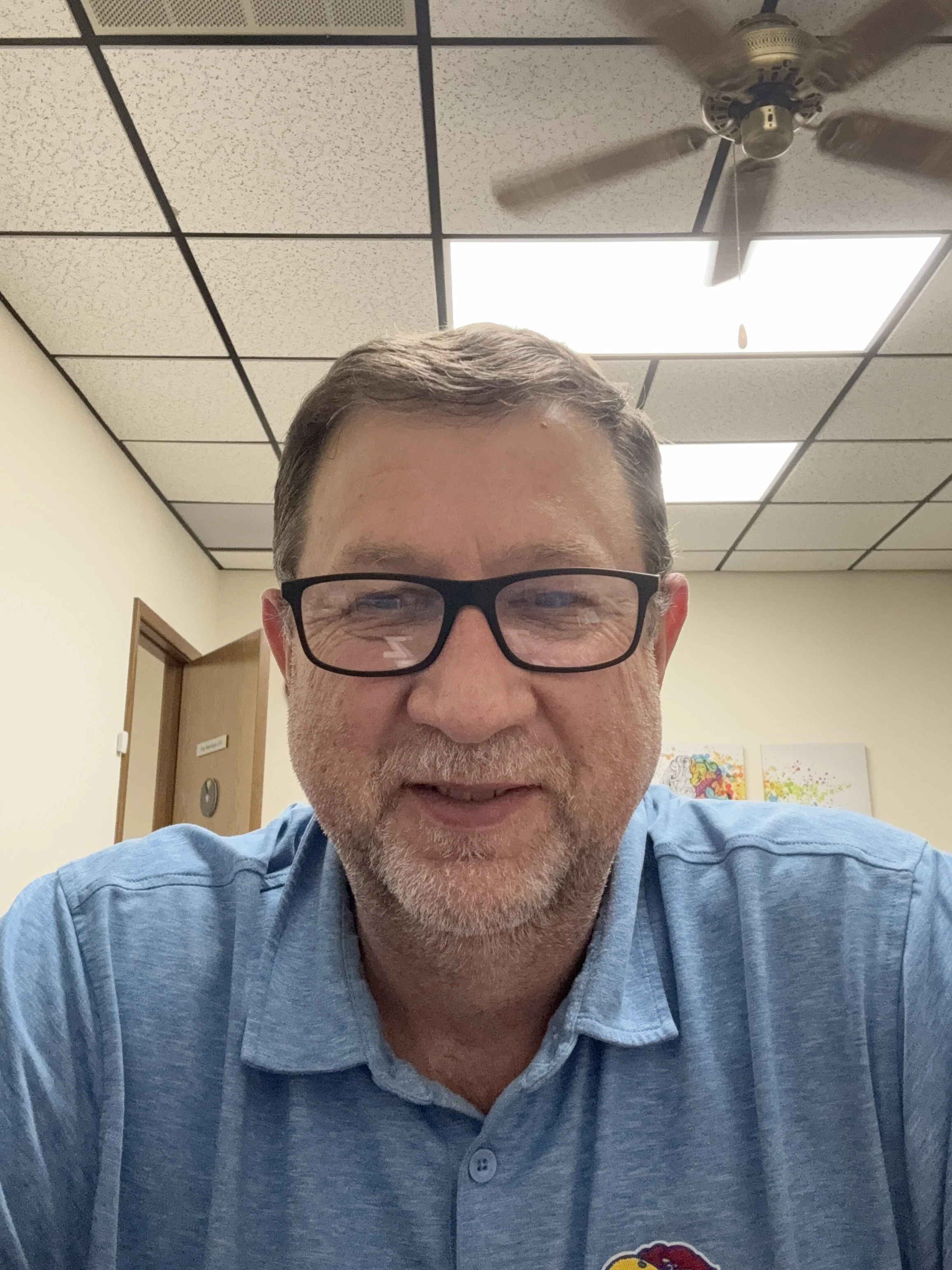 A middle-aged man with glasses, gray hair, and a beard is smiling in a close-up selfie. He is wearing a blue collared shirt with a University of Kansas logo. The background shows a ceiling with fluorescent lights, a ceiling fan, a wooden door, and colorful artwork on the wall.