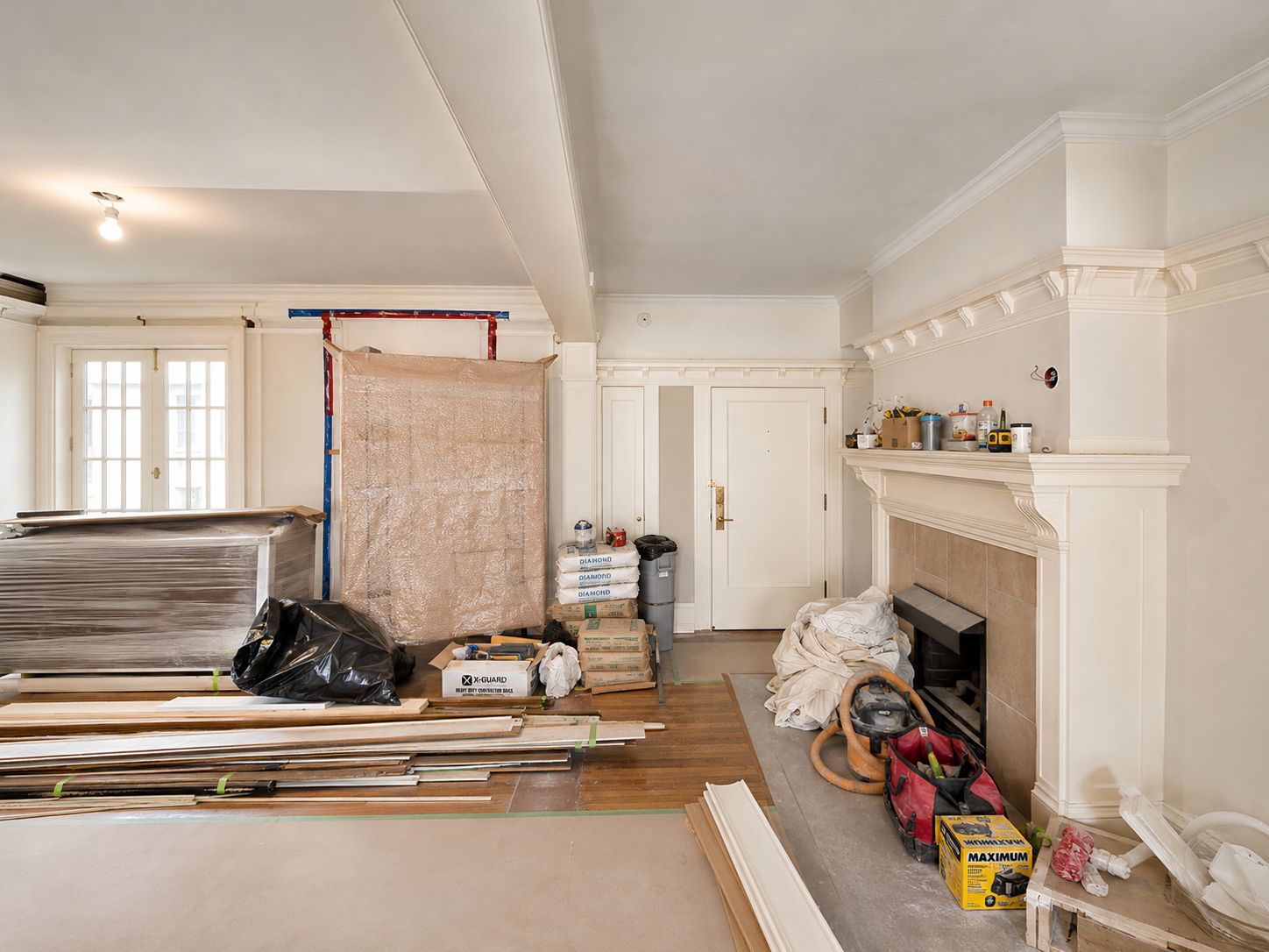 Living room under renovation with construction tools, materials, and equipment scattered around, including wood, drywall, and paint supplies.