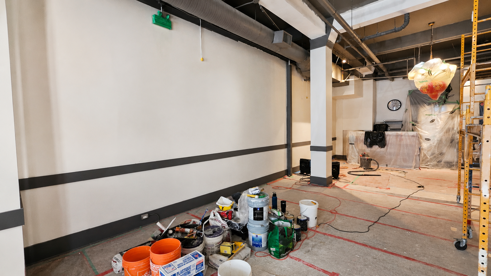 An interior room under renovation with construction materials, tools, buckets, and equipment scattered on the floor. Plastic sheeting covers furniture and fixtures, and there are exposed pipes and a ladder.